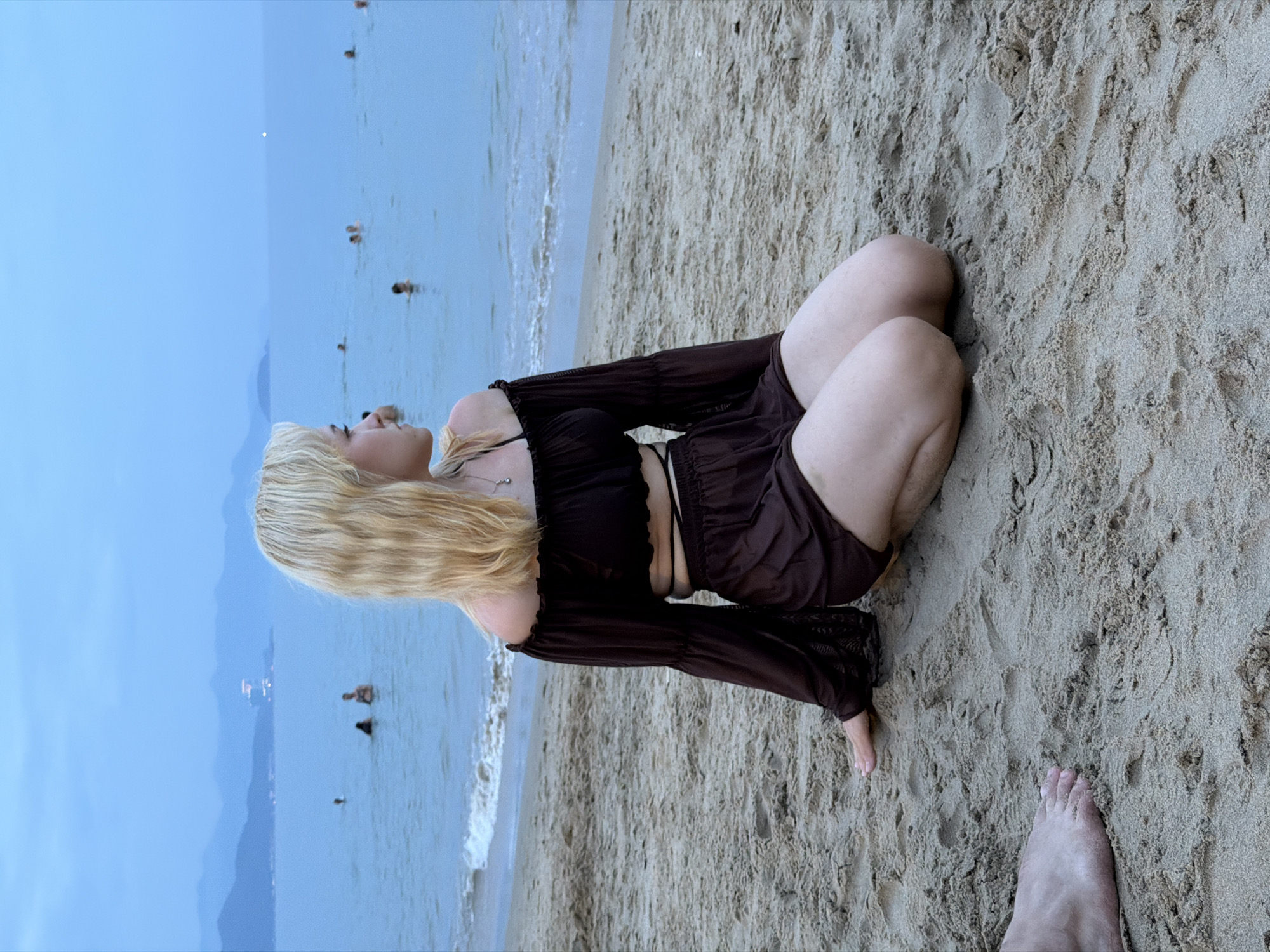 The image depicts a young Caucasian woman with blonde hair kneeling on a sandy beach, facing away from the camera and looking out at the ocean. She is wearing a dark brown, off-the-shoulder top with bell sleeves and matching shorts. Her left hand is resting on the sand, and her knees are bent. In the background, the ocean is calm with a few people swimming. The sky is overcast and a dusky blue, with a hazy mountainous coastline visible in the distance. The scene is characteristic of a beach in Nha Trang, Vietnam, likely during the evening or early morning hours due to the soft, diffused light and the muted colors. The presence of the ocean, sand, and distant mountains are consistent with the coastal geography of Nha Trang. A bare foot is visible in the lower left corner of the frame, suggesting another person is present just outside the main subject's view.