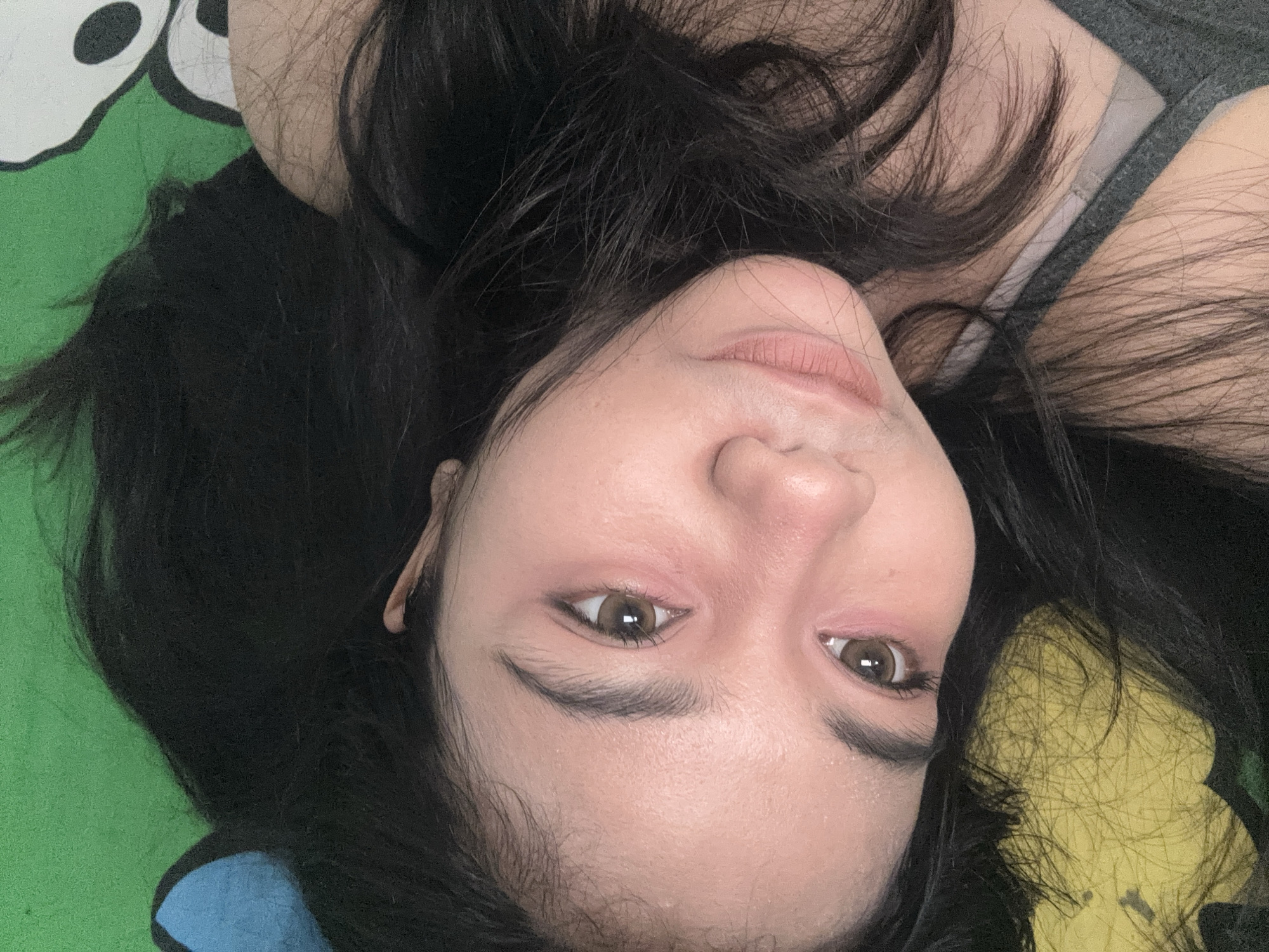 The image shows a person lying on a colorful, patterned surface. The person has dark hair and light skin, with visible makeup around their eyes. They are wearing a gray top, and the background features a green, yellow, and blue pattern. The location cannot be determined from this image.