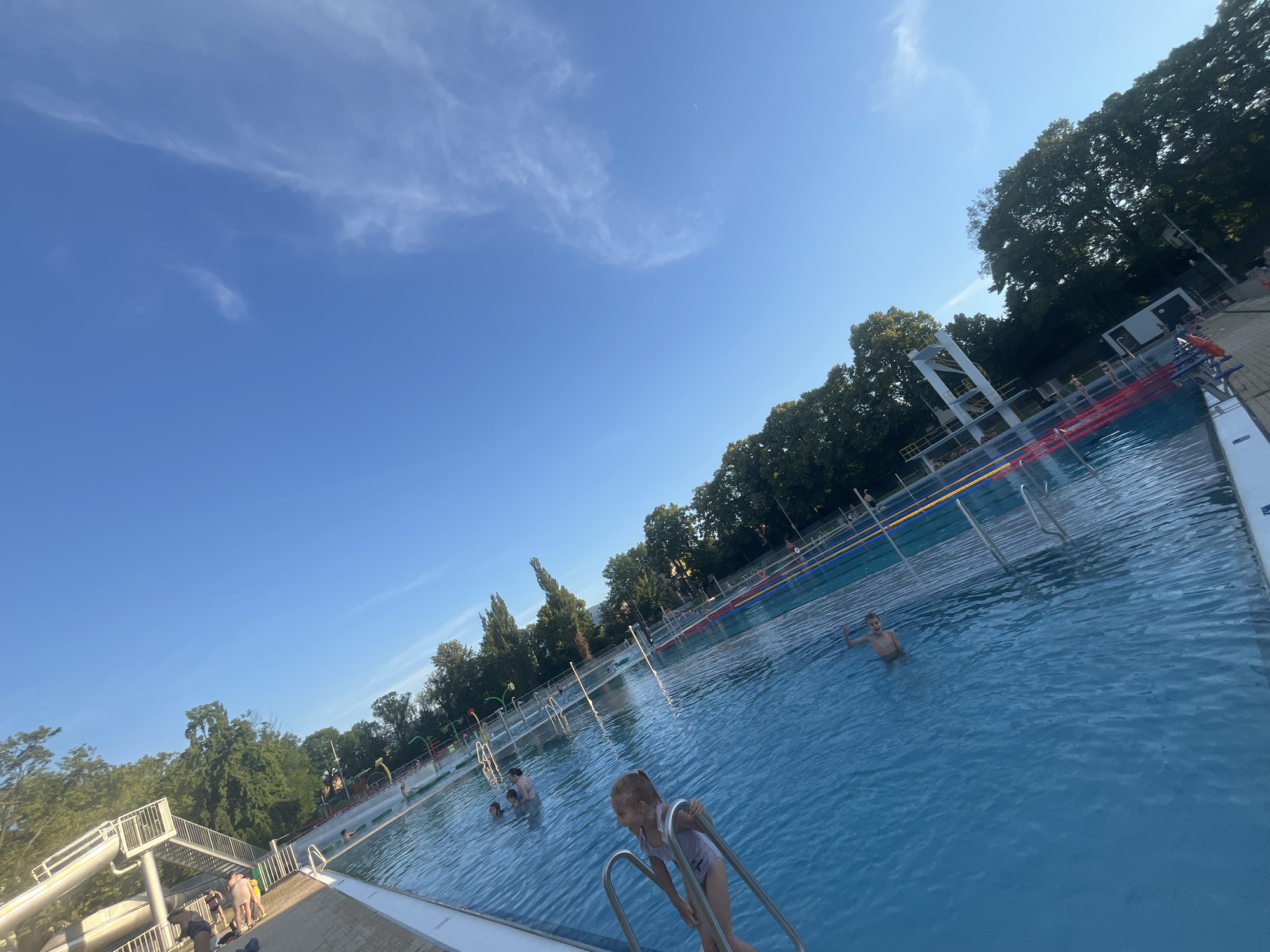 The image shows a bright blue swimming pool on a sunny day, with a clear blue sky and scattered white clouds. The pool is divided into lanes with colored markers. There are several people in the pool, including children and adults, some of whom are swimming or playing. On the right side of the pool, there are several lane ropes and a diving platform with a white structure. Trees and buildings are visible in the background. In the foreground, a young girl is holding onto the ladder of the pool. The image is taken from a low angle, with the pool occupying most of the frame.