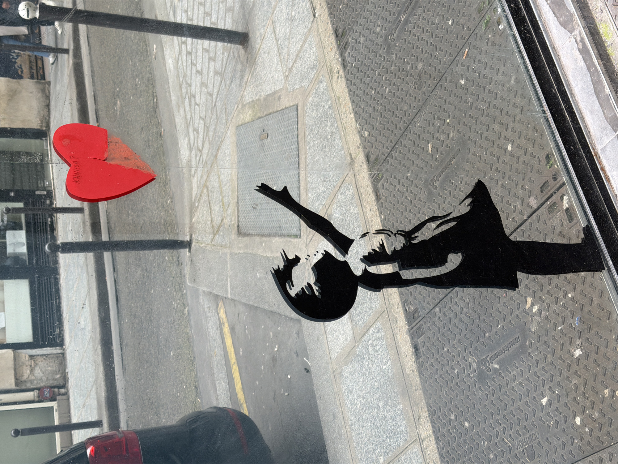 The image shows a Banksy-style artwork on glass, seemingly a window display or installation. The artwork depicts a black silhouette of a person reaching for a red heart-shaped object resembling a balloon. The red heart is partially damaged, appearing broken. The background reflected in the glass shows a Parisian street scene, including pavement, a street lamp, and a building. A car is also visible in the reflection at the bottom. The manhole cover indicates that the location is most likely in Paris, France.