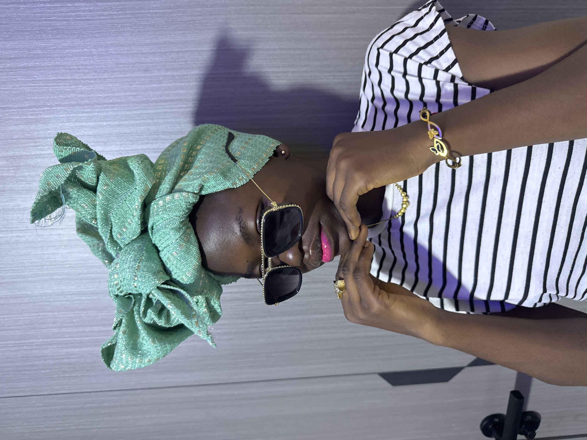 A person with dark skin is wearing a light green headwrap, black sunglasses, a striped black and white shirt, and gold jewelry. The person is posing indoors against a light gray wall. A camera tripod can be seen in the lower right corner. The location is unknown, and no evidence points to Ajara, Nigeria.