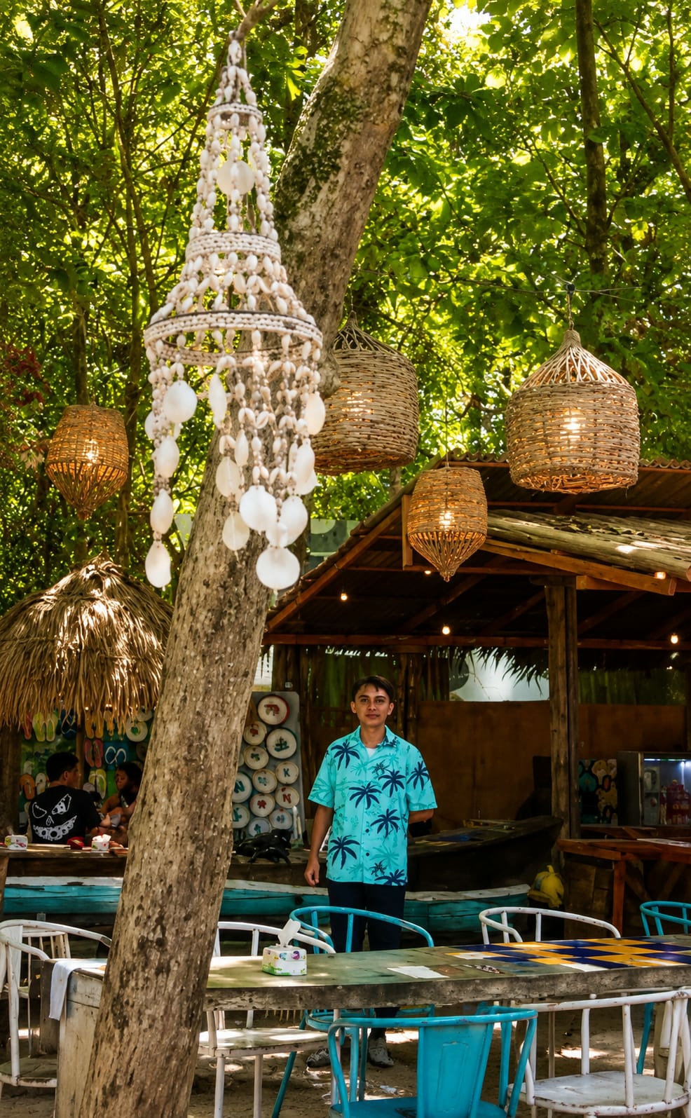 A young man wearing a light blue short-sleeved shirt with a palm tree pattern and dark pants stands in front of a rustic open-air establishment. The establishment features a thatched roof and is decorated with woven pendant lights and a shell chandelier. Tables and chairs are set up in the foreground, some made of wood and painted with turquoise accents. In the background, there are other diners seated at a table, and a display of colorful flip-flops. The scene is set outdoors amidst lush green trees, suggesting a tropical or coastal location. The overall atmosphere is casual and relaxed, indicative of a beachside cafe or restaurant. The city of Medan, Indonesia is implied as the location by the user.