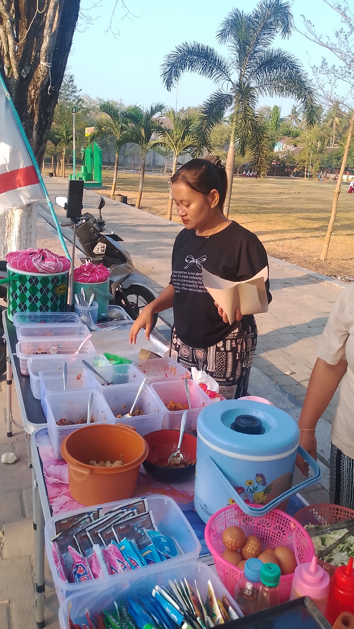 A woman stands behind a table of food items outdoors in Karanglangu, Indonesia. She is wearing a black t-shirt with a white graphic and patterned pants. In her right hand, she holds a beige paper cone. She is looking down at the food on the table. The table is covered with a colorful cloth and holds various containers of food, plastic bags of powdered drinks, a basket of eggs, and condiments. To the left of the table, a motorcycle is parked. In the background, palm trees and a clear sky suggest a warm climate.