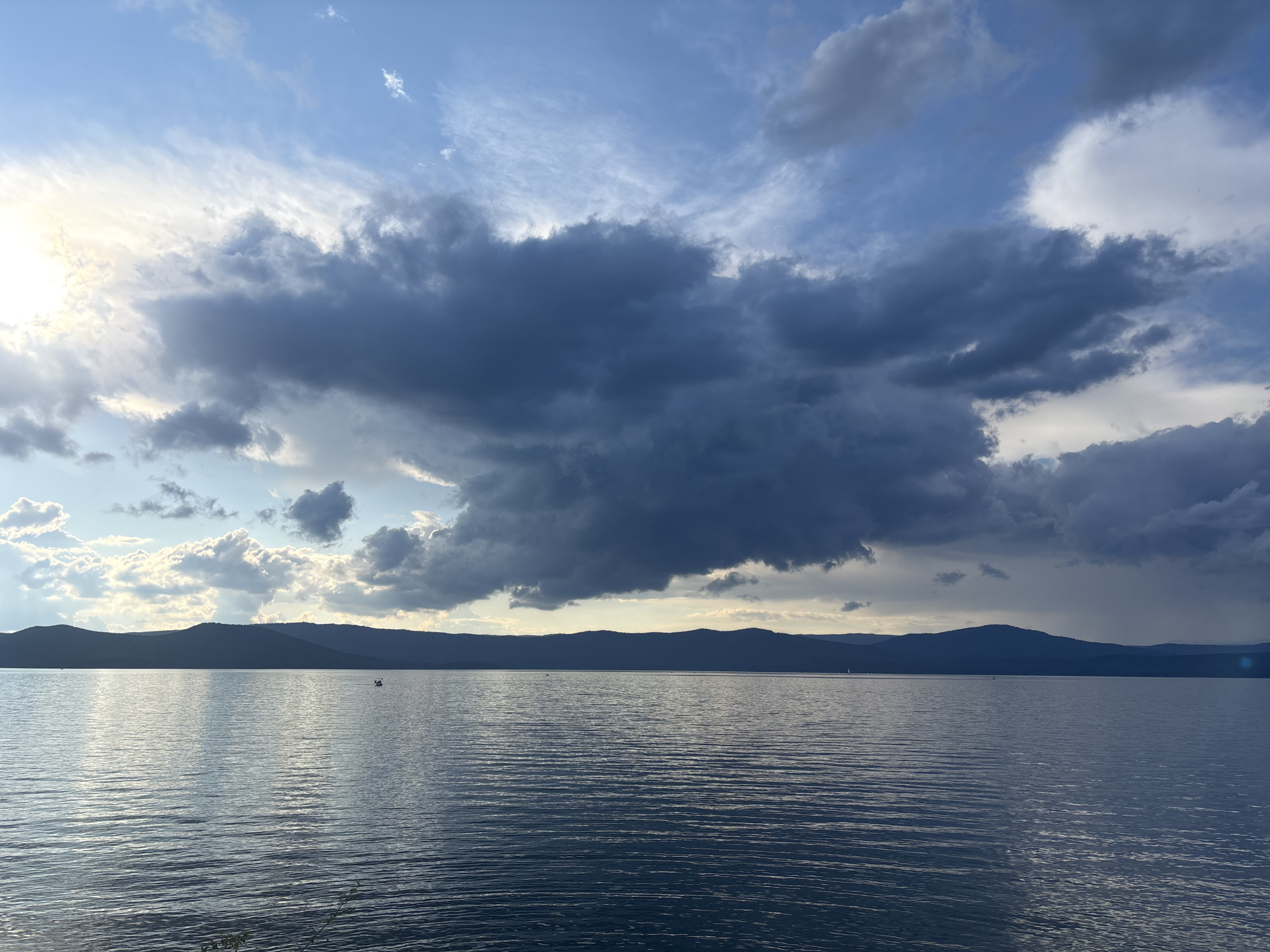 An outdoor, wide shot captures a serene, large body of water at dusk. The water, reflecting the sky, is calm with a slight ripple. In the distance, a dark, silhouetted mountain range stretches across the horizon. The sky is a dramatic mix of blues and grays, with heavy, dark clouds dominating the center and lighter clouds and a bright spot of sunlight on the left. A small boat is visible on the water, adding a sense of scale. The location is Turgoyak, Russia, a resort town known for its clear lake.