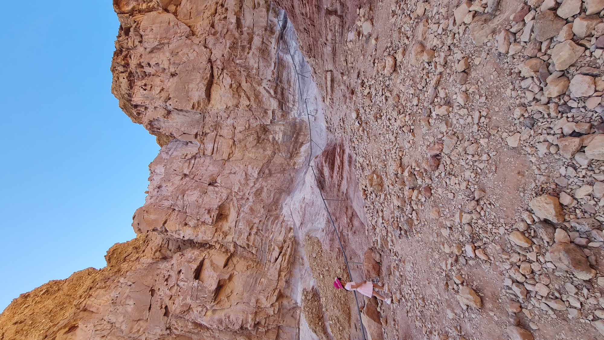 A young girl in a pink dress and hat stands on a rocky, uneven terrain at the base of a steep rock cliff face. The cliff, composed of reddish-brown stone, is marked with a series of metal bars that form a rudimentary ladder system ascending the rock face. The sky is visible above the cliff. This scene is located in the city of Hevel Eilot Regional Council, Israel.
