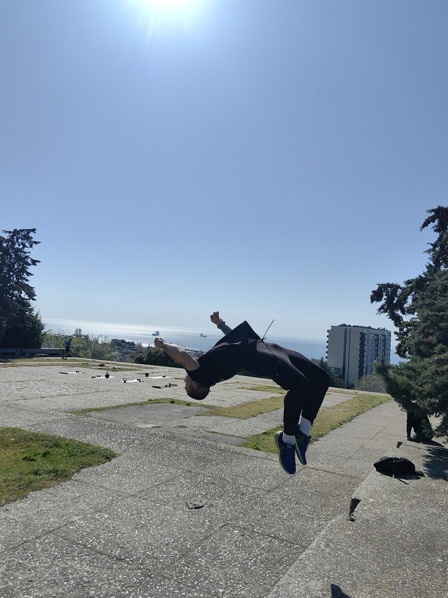 The image shows a person performing a backflip in an outdoor location. The person is wearing dark clothing and blue sneakers with white socks. The background includes a body of water, a cityscape, trees, and a paved area. There is fitness equipment such as mats and kettlebells on the paved area. A person is running in the background. There is a bag on the ground. The sky is blue. The location is in the city of Unknown, Unknown.