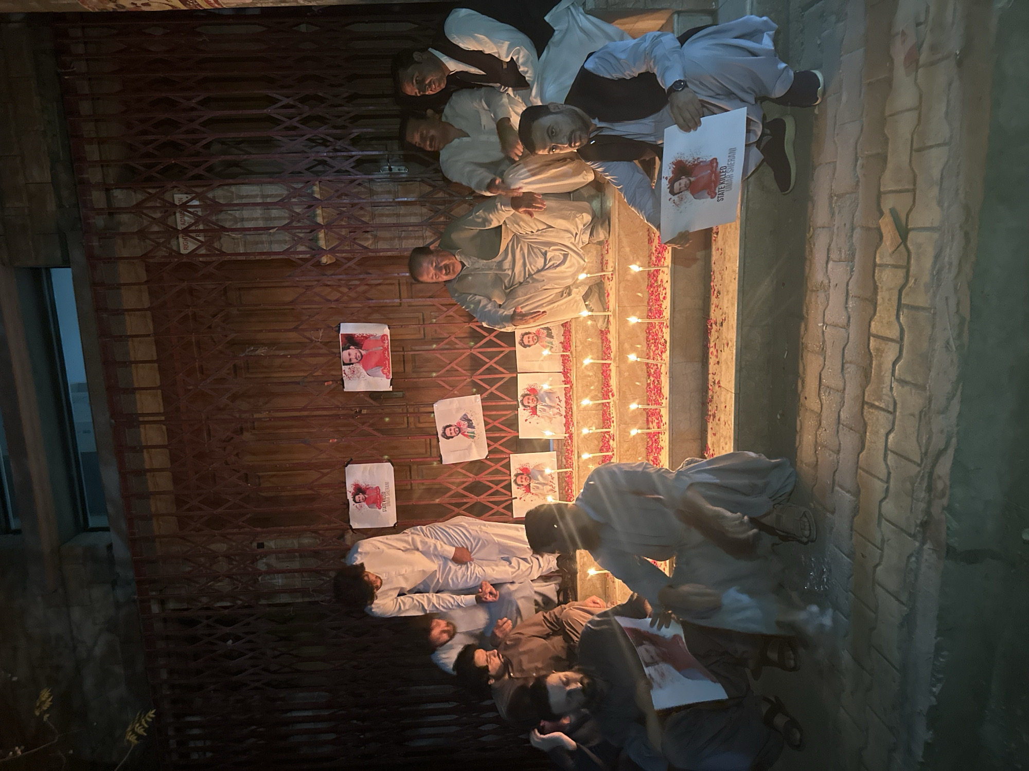 In Quetta City Tehsil, Pakistan, a group of individuals is gathered in front of a building with a red metal security gate. The scene is lit by numerous lit candles arranged on steps and scattered with red petals. Several people, dressed in traditional Pakistani attire, are seated or crouching on the steps and pavement. Some are holding up printed photographs of a man. These photographs appear to be memorials, with some bearing text that reads "STATE KILLED UMAR SHERANI." The overall atmosphere suggests a vigil or memorial service. The background shows the dark facade of the building and the metallic grating of the security gate.