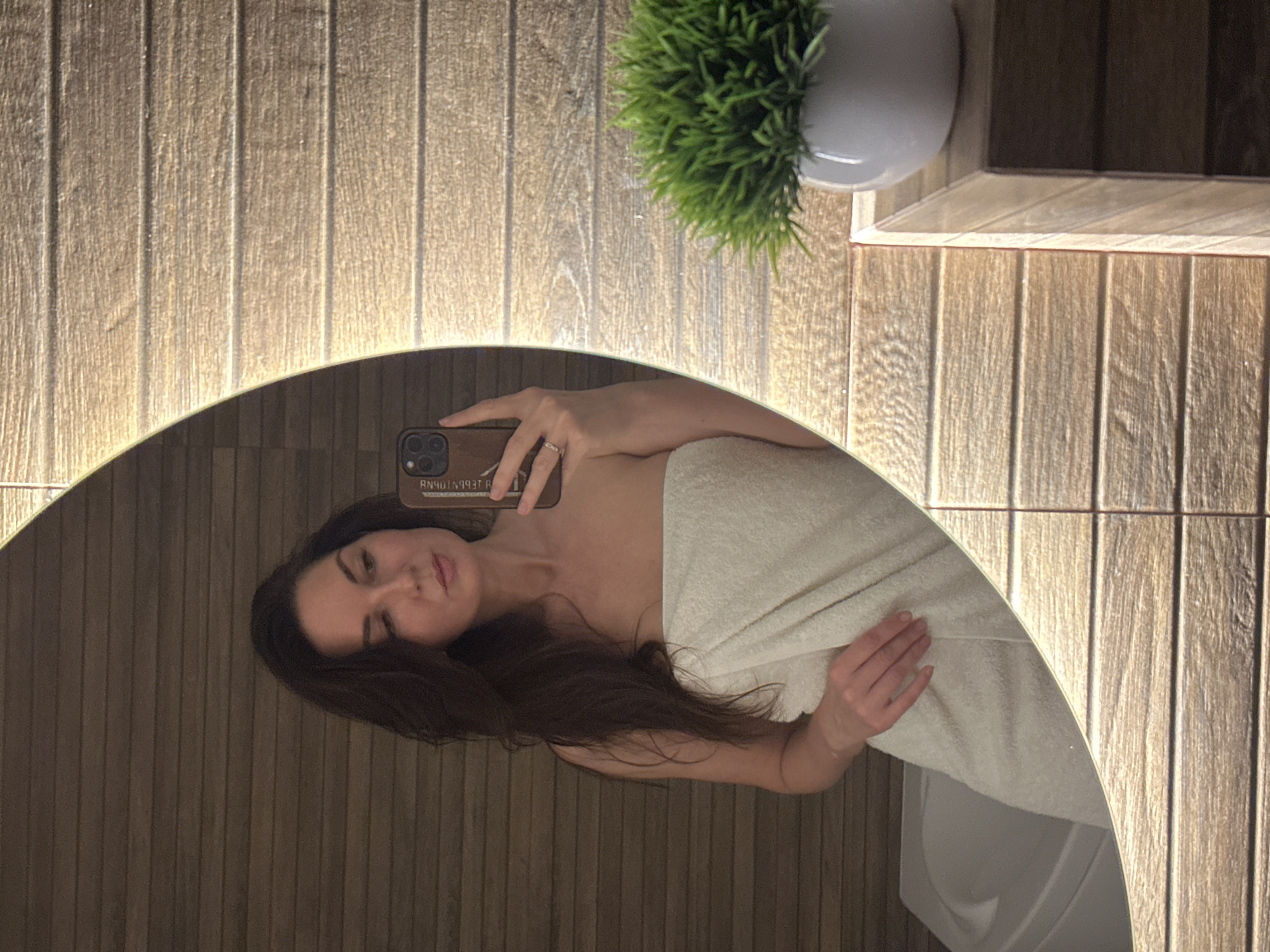 A woman wrapped in a white towel is reflected in a round, illuminated mirror. She is holding a smartphone to take a selfie. The background consists of vertical wooden paneling with light fixtures between the panels. A potted green plant is on a shelf in the upper right corner. The location and city of Chaikovsky, Russia are not possible to ascertain from the image.