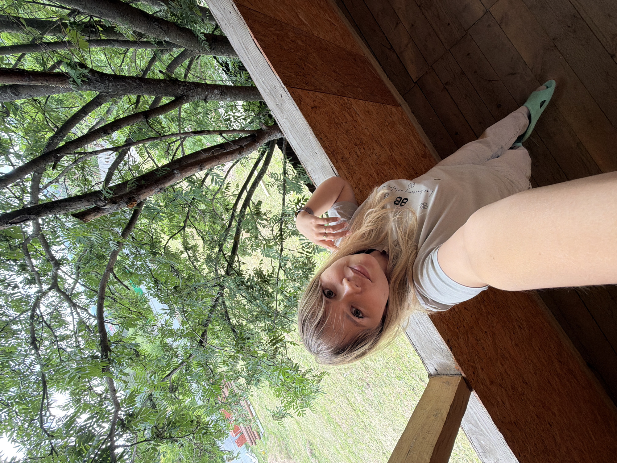 An image, rotated 90 degrees clockwise, features a young woman posing for a photo on an elevated wooden structure. She has long, blonde hair with bangs and is looking directly at the camera with a calm, slightly smiling expression. She is wearing a light gray t-shirt with partially visible black text that reads "88" and "better love, better life...", along with light-colored pants and light mint-green slip-on shoes. Rings are visible on her left hand.
The wooden structure consists of rustic plywood paneling and sturdy beams, suggesting a porch, balcony, or similar outdoor platform. To her left, a dense canopy of green tree branches and leaves fills a large portion of the background, indicating close proximity to lush foliage. Patches of green grass are visible below the trees, suggesting an outdoor, natural environment. The scene is brightly lit, indicating daytime. The context places the location in Chaikovsky, Russia.
