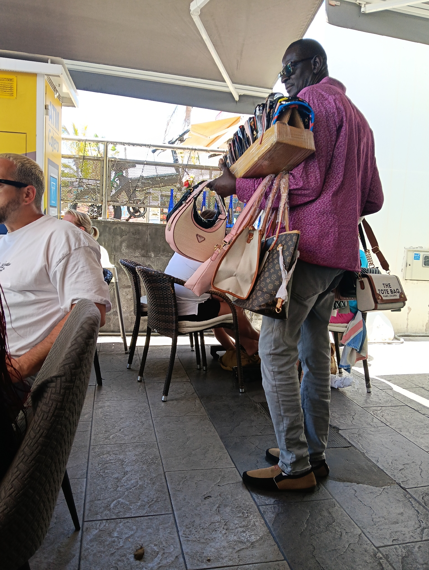 The image depicts an outdoor daytime scene in what appears to be a cafe or public area in Puerto Del Carmen, Spain. In the foreground, a Black man, likely a vendor, is seen in profile, walking or standing. He wears a patterned purple long-sleeved shirt, grey jeans, and two-toned tan and black slip-on shoes. He carries a wooden rack laden with multiple pairs of sunglasses on his right shoulder, while several handbags hang from his left arm and shoulder.
Among the handbags are a light pink and beige bag with "PRADA" visible on it, a brown bag with a distinct monogram pattern resembling Louis Vuitton, and a beige canvas tote bag clearly marked "THE TOTE BAG MARC JACOBS".
To the left of the vendor, two customers are seated at cafe tables. The closer customer is a man with short blonde hair and sunglasses, wearing a white T-shirt, visible from the chest up. Another person is partially visible sitting further back. The ground is paved with large, textured grey tiles.
In the background, a yellow structure with the text "ATM" and "Cajero" (Spanish for ATM) is visible, indicating a commercial zone. The bright lighting and shadows suggest a sunny day. The scene captures a casual public interaction, with a vendor approaching or passing by seated customers.