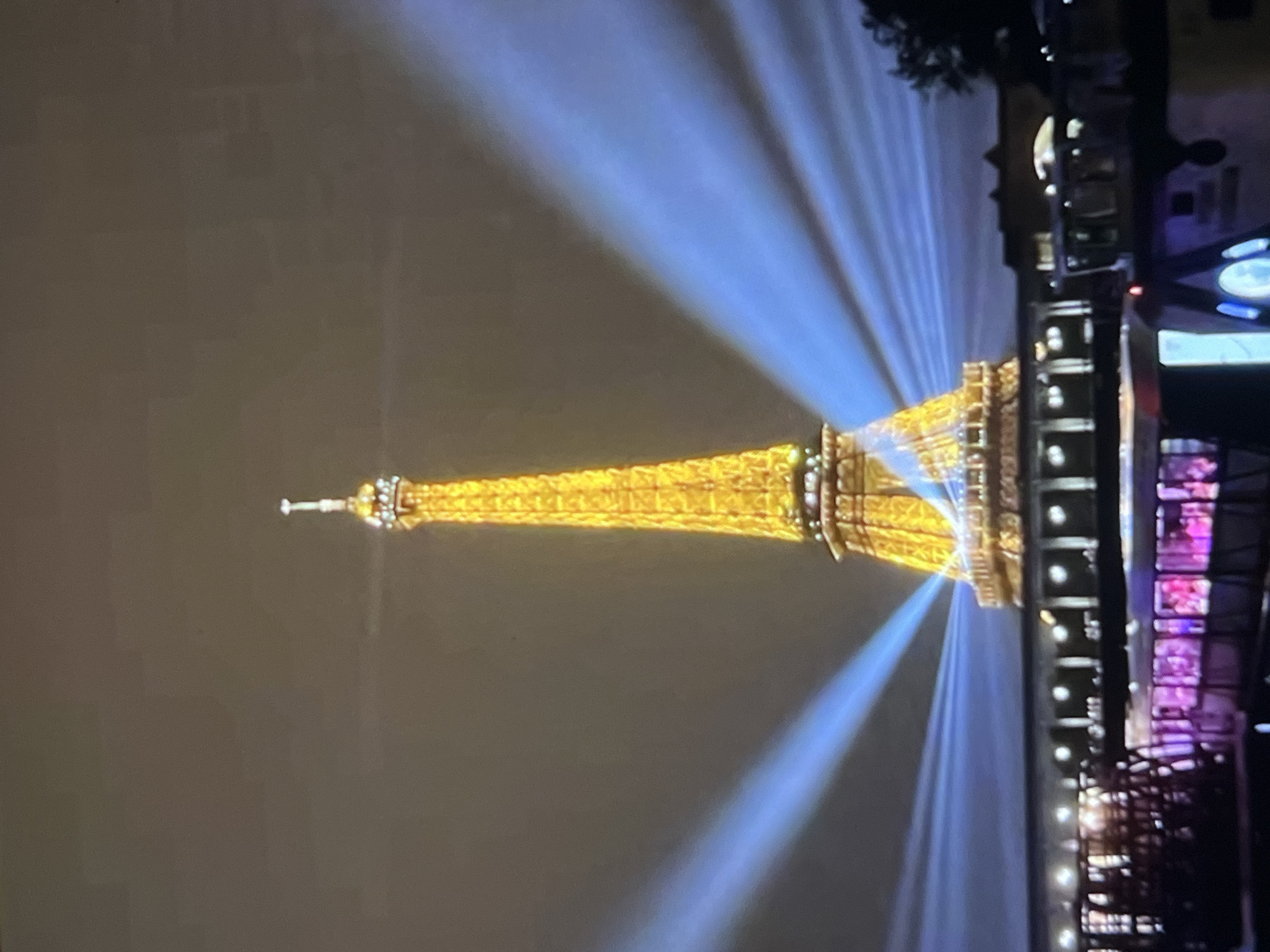 The image shows the Eiffel Tower illuminated with golden lights at night, with bright blue beams of light emanating from its base and reaching towards the sky. The tower is situated in the city of Paris, France, not Montfermeil. In the foreground, there is a body of water with several boats, one of which has colorful lights. A structure resembling a bridge or promenade runs along the water's edge in front of the tower, with lights illuminating it. The sky is dark and overcast.