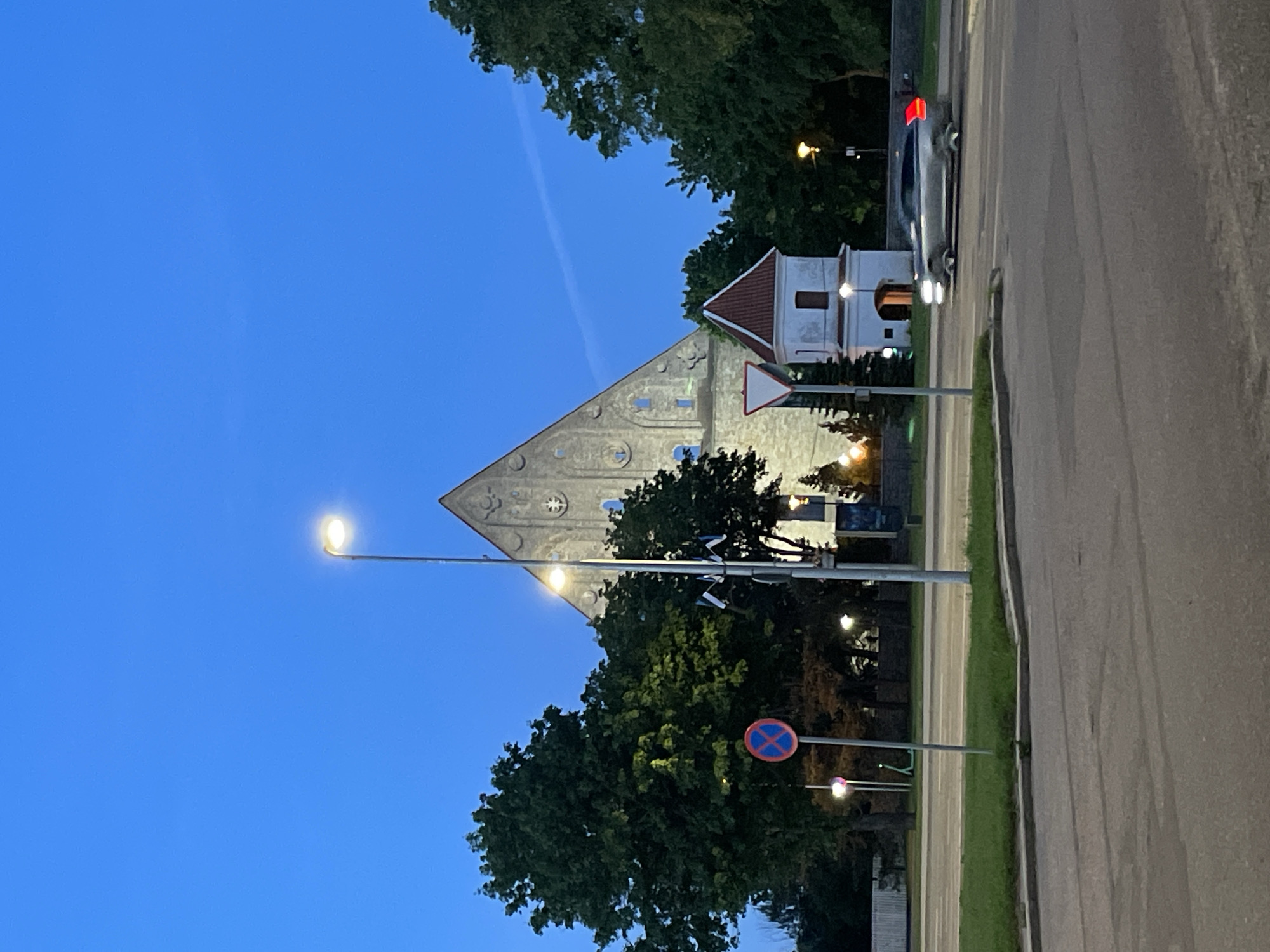 The image captures a twilight scene in Tallinn, Estonia, focusing on the Kaarli Church, a neo-Gothic Lutheran church with a distinctive triangular roof. The sky is a deep blue, indicating dusk.
In the foreground, a gray car is turning right at an intersection. The car's headlights and taillights are illuminated, suggesting it's in motion. A traffic light with a red signal is visible on the left side of the image, near a circular red and blue sign indicating a restriction. A street lamp, also lit, stands in the middle ground.
Behind the intersection and street lamp, the imposing structure of the Kaarli Church dominates the background. Its light-colored stone facade is illuminated by interior lighting, highlighting its windows and architectural details. To the right of the church, a smaller, white building with a red roof and arched entryway is also visible.
Several trees with lush green foliage surround the church and the smaller building, their shapes softened by the low light. Two Estonian flags are mounted on a pole near the church. The overall atmosphere is serene, with the artificial lights of the city beginning to counter the fading natural light. The scene suggests a typical urban environment at the transition from day to night.