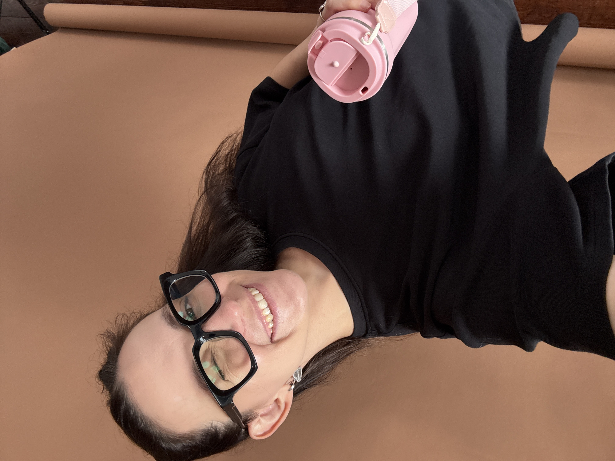 A woman with dark hair and glasses smiles while looking at the camera. She is wearing a black t-shirt and holding a pink water bottle in her right hand. The background is a solid brown color. The image appears to be a selfie taken indoors. There is no discernible location in Chaikovsky, Russia, indicated in the image.