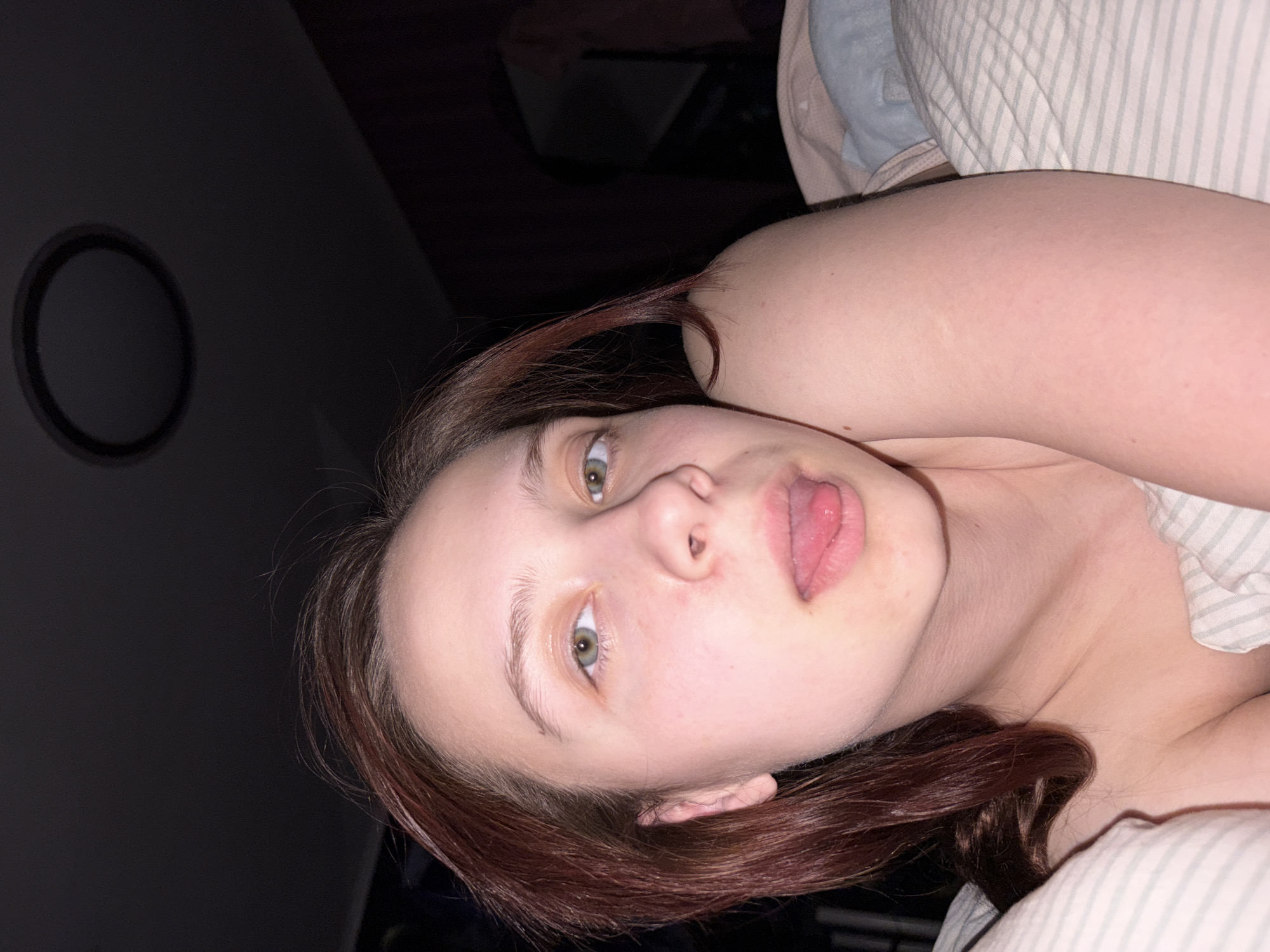 A woman with fair skin and reddish-brown hair is lying on a bed. Her face is turned towards the camera with her tongue sticking out. She has light-colored eyes. The background is dark with a circular light fixture visible on the wall. The setting appears to be indoors. The location is Прикамский, Russia.