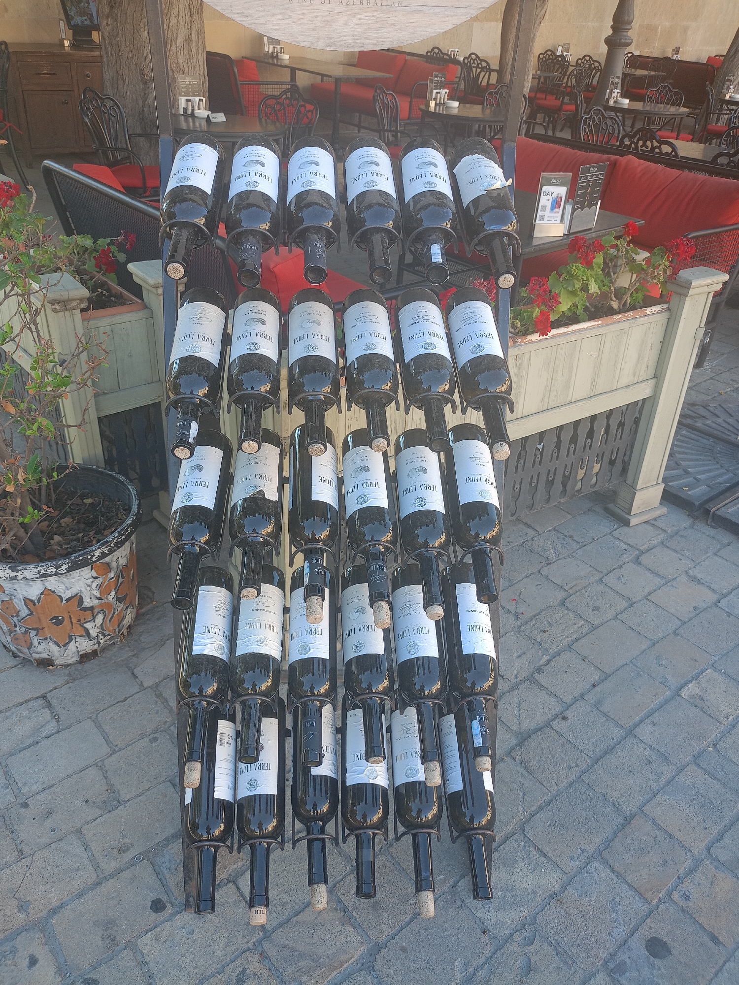 The image shows a display of wine bottles on a rack in an outdoor seating area of a restaurant or cafe. The bottles are dark glass and have white labels. The rack is made of metal and is positioned on a paved walkway. In the background, there are tables and chairs with red upholstery, suggesting an outdoor dining space. There are also potted plants and a tree trunk visible. The overall scene is one of a well-maintained establishment, likely in a European city given the architectural style.