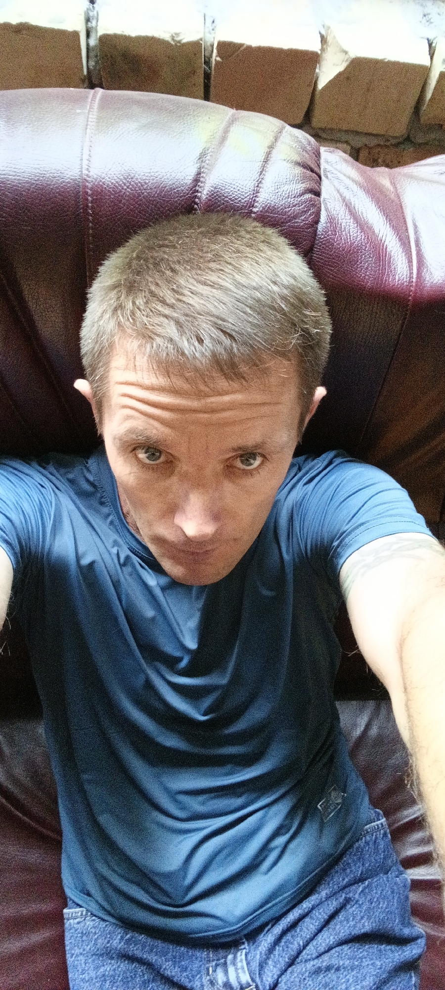 The image is a close-up, high-angle selfie of a man sitting on a dark red leather sofa. He has short, light brown hair and is looking directly at the camera with a serious expression. He is wearing a blue t-shirt and blue jeans. The background shows a brick wall with horizontal lines. The scene does not provide any information about the location in Tashkent, Uzbekistan.