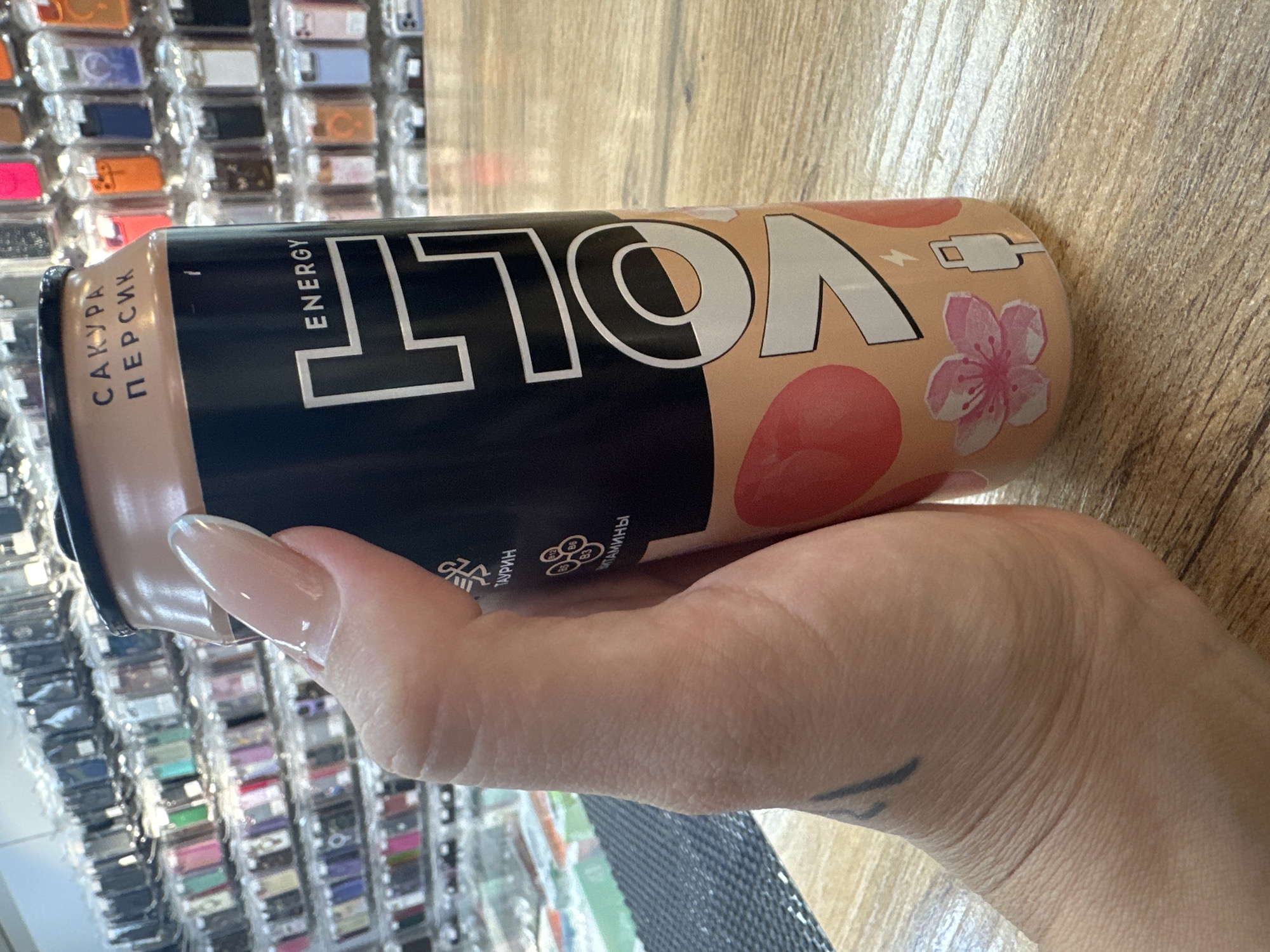 The image shows a close-up of a person's hand holding a can of "Volt Energy" drink with "Sakura Peach" flavor. The can is black and peach-colored, with white text and graphics including cherry blossoms and a USB symbol. In the background, shelves filled with phone cases are visible. The setting appears to be a store, possibly a mobile phone accessory shop. The wooden surface the can is resting on is also visible. The specific location in Partisansk, Russia is not discernible from the image itself, but it is implied by the prompt.