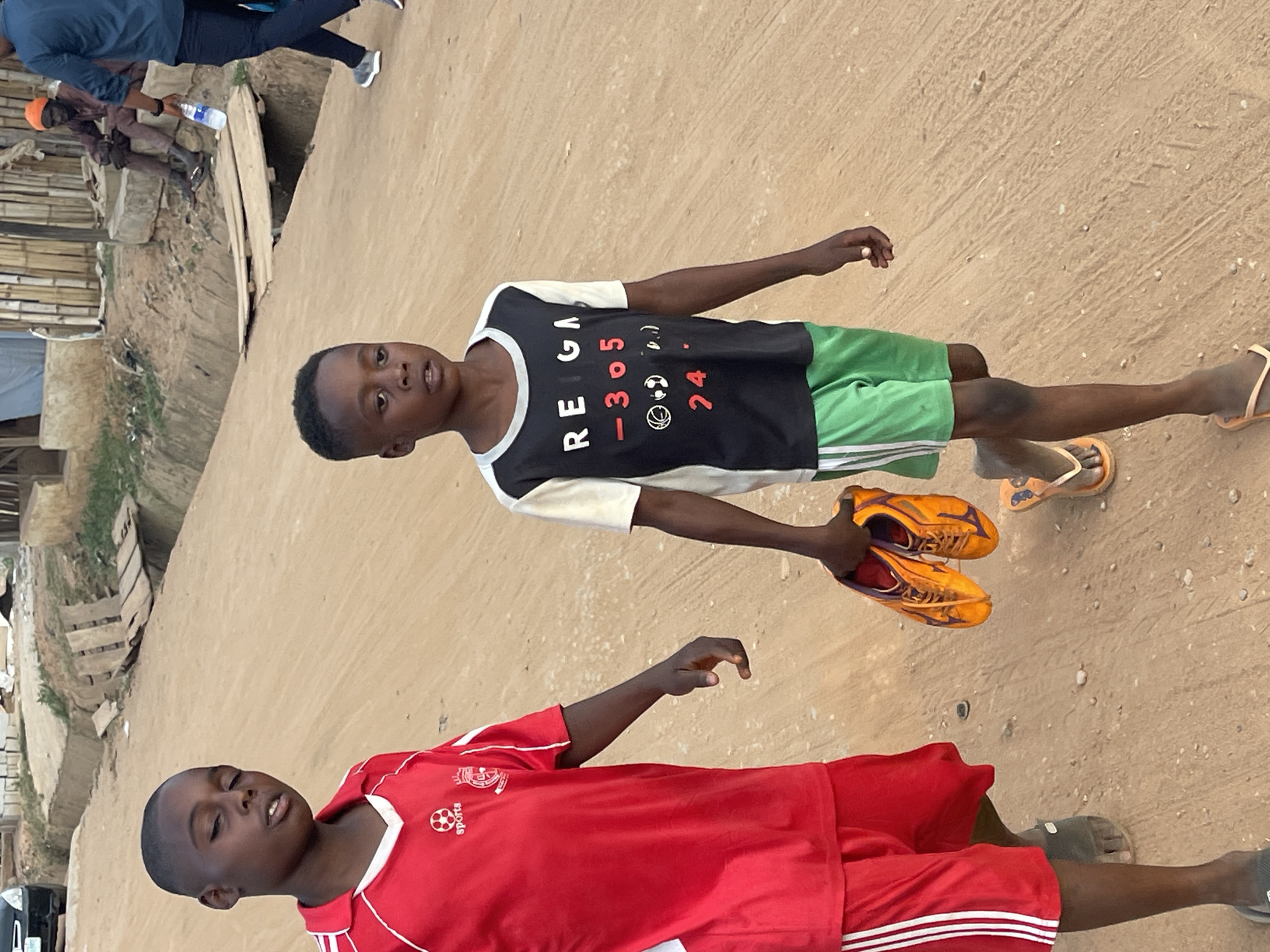 Two African boys stand on a dirt road. One boy is wearing a black shirt with a white design and green shorts, carrying orange soccer cleats. The other boy wears a red soccer jersey and shorts with white stripes. The setting appears to be Karmajiji, Nigeria, judging by the visible, partially-constructed buildings and infrastructure in the background, along with clothing styles and facial features of the boys. Additional people are seen on the left side of the image, further in the distance.