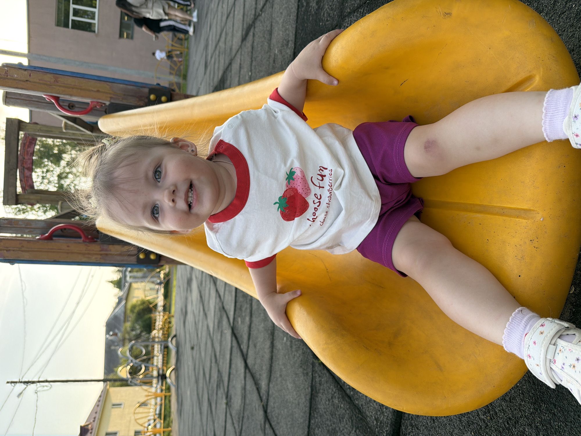 A young Caucasian girl with blonde hair and blue eyes is positioned on a yellow playground slide. She wears a white t-shirt with red trim and a strawberry design, purple shorts, white socks with a floral pattern, and white sandals. The girl is looking at the camera with a smile. The playground is located outdoors. The background includes buildings, trees, and playground equipment. Reshetnikovo, Russia, is the city location.