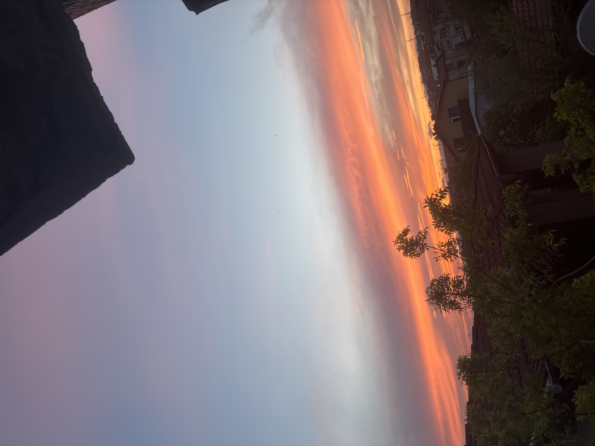 The image captures a view from Burgas, Bulgaria during sunset. A portion of a building or roof is visible in the upper left corner, providing context for the viewpoint. The sky displays a gradient, transitioning from shades of pink and purple near the bottom to hues of orange and yellow toward the horizon, indicative of sunset colors. In the distance, buildings and structures are silhouetted against the bright sky. Branches and foliage are present in the foreground along the right side of the frame, adding depth and a sense of urban nature. There are no people visible in the image.