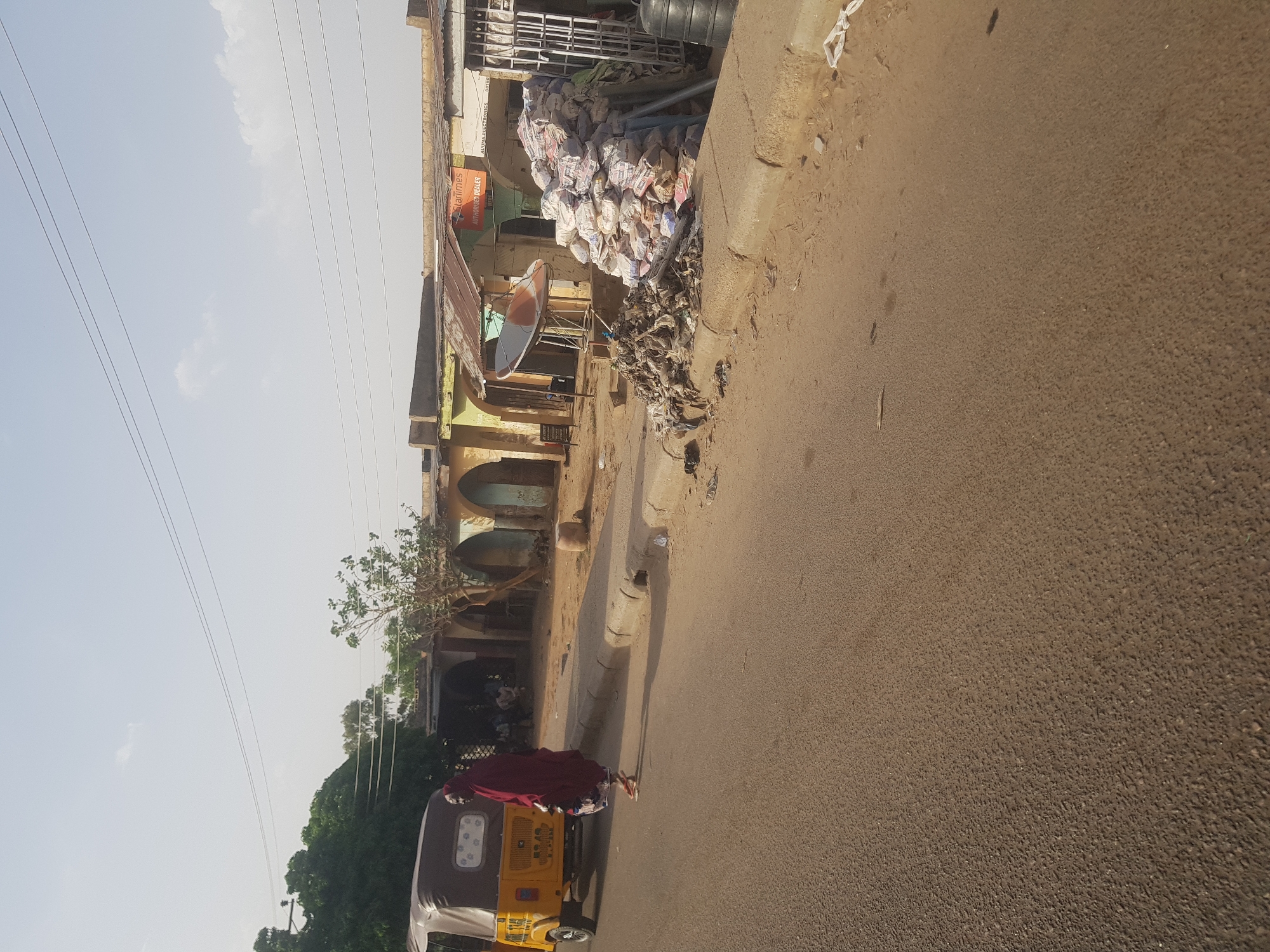 A medium shot captures a street scene in Dutsinma A, Nigeria, featuring a brown and yellow Bajaj tricycle, commonly known as a "Keke," on the left side of the asphalt road. A woman wearing a red head covering and gown is positioned in front of the vehicle. Stacks of white sacks are piled on the side of the road near a multi-story building. The building has a faded yellow façade, a green arched colonnade, and a satellite dish attached to the upper part of the building. Wires are stretched across the sky on the left side of the image.