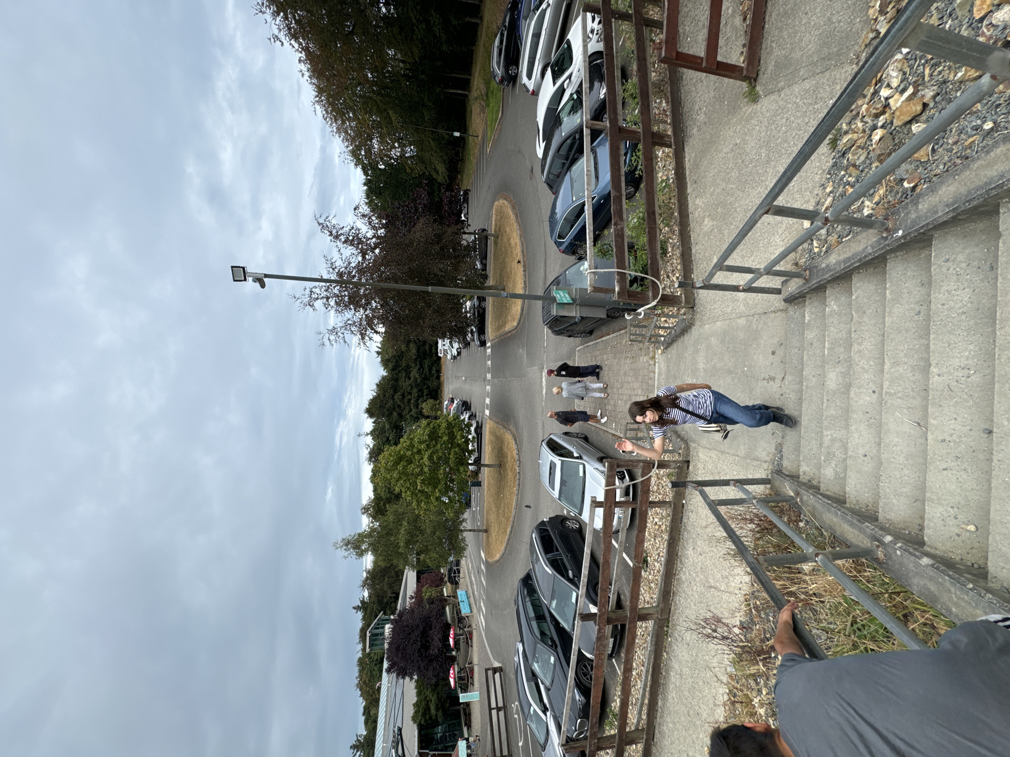 The image depicts an outdoor scene with a parking lot and a building in the background, likely a visitor center or restaurant. A young woman in a striped shirt and jeans is walking up a set of concrete stairs, while a man in a grey t-shirt is descending the stairs. Several cars are parked in the lot. The environment appears to be a natural setting with trees and foliage. The sky is overcast. The location is identified as Courtown, Ireland.