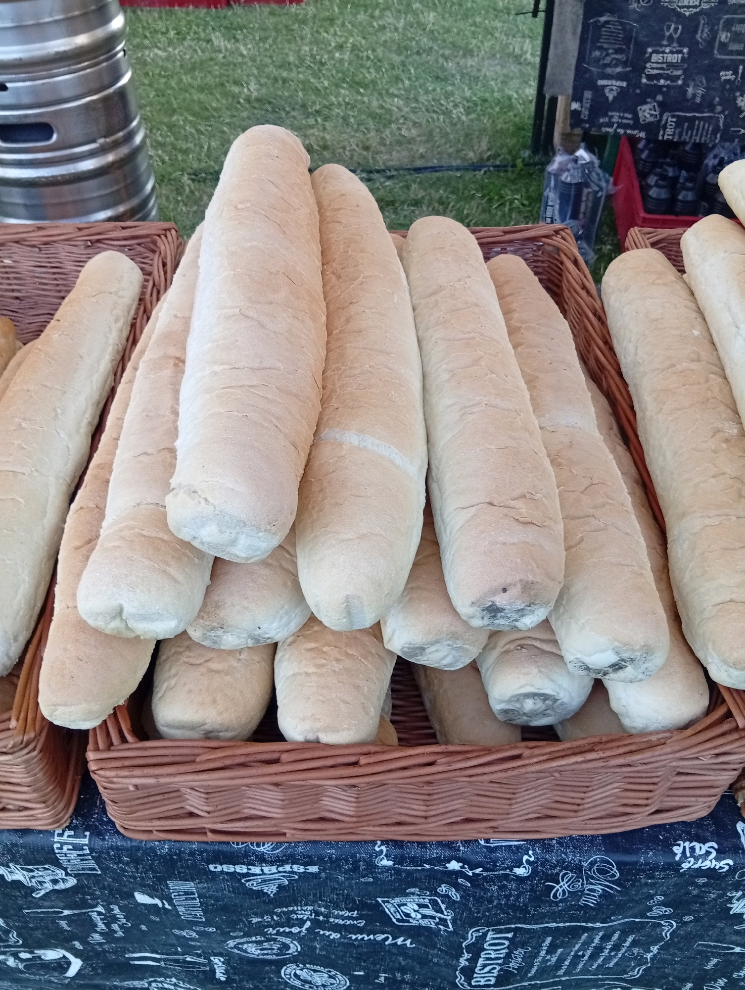 A close-up, high-angle outdoor shot shows several loaves of bread arranged in brown woven baskets, placed on a dark fabric tablecloth with white lettering and graphics. The bread loaves are long and slender, with a light brown crust and a slightly textured surface. The baskets are positioned on a table, with the tablecloth covering the surface beneath. In the background, a silver metallic barrel or keg is visible to the left, and a grassy area and a blackboard sign can be seen to the right. The scene appears to be set up at an outdoor market or food stall.