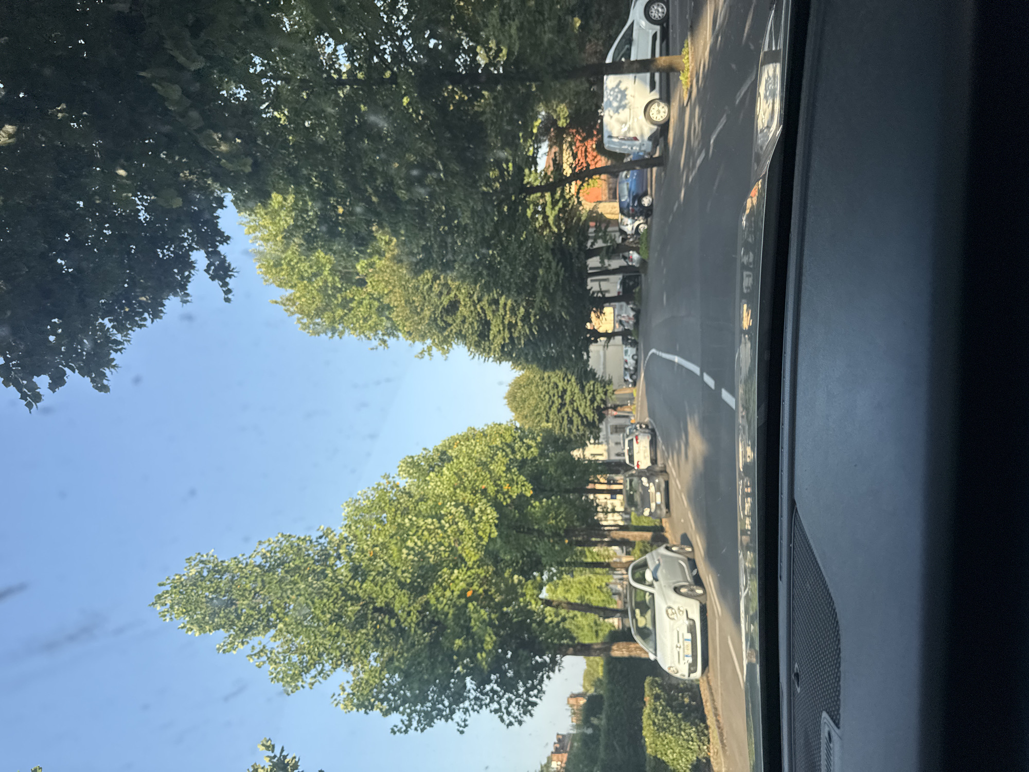 The image depicts a street scene in Chiari, Italy, viewed from the interior of a vehicle. Tall, mature trees line both sides of a paved road with white lane markings. Several cars are parked along the street and on the roadside. On the left, a white Fiat 500 is prominently parked. Further down the street, other vehicles, including a dark hatchback and a white van, are visible. The sky is clear and blue, suggesting daylight. The presence of lush green foliage indicates a temperate season. The overall impression is that of a quiet residential or commercial street.