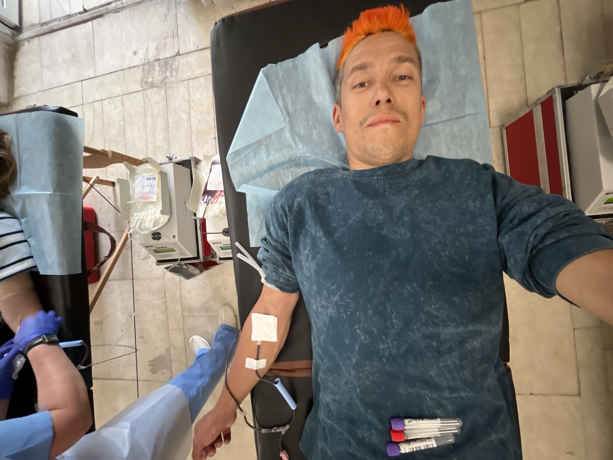 A first-person view shows a person lying on a medical examination table, presumably donating blood. The person has orange hair and is wearing a blue shirt. A medical professional is drawing blood from the person's arm. Blood tubes and collection equipment are visible. The location is a medical facility. The specific location in Chaikovsky, Russia, cannot be determined from the image.