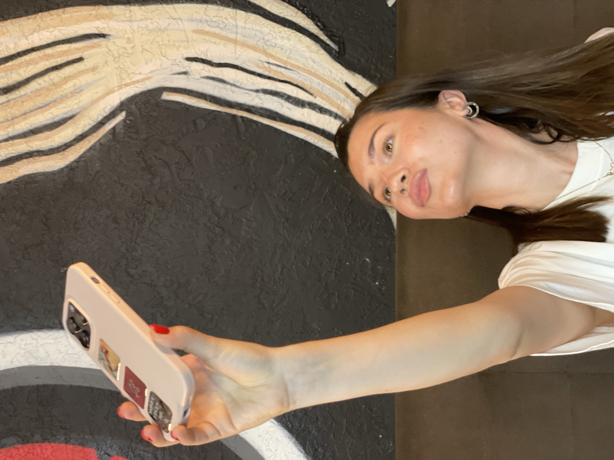 A young woman is taking a selfie with her mobile phone. She has long brown hair, light skin, and is wearing hoop earrings. She is wearing a white t-shirt and has a necklace. Her fingernails are painted red. The phone has a light pink case with stickers on the back. The background features a wall with black, white, and beige abstract art. The location is identified as Krasnoyarsk, Russia.