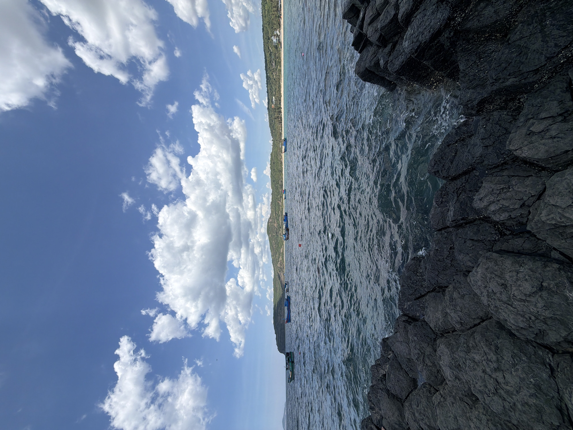 This media file depicts a coastal scene in Phú Lương, Vietnam. The composition is dominated by a dramatic contrast between a deep blue sky filled with fluffy white clouds and the dark, textured surface of volcanic rocks in the foreground. To the right of the frame, these rocks plunge into the dark, rippling water of the sea.
Stretching across the middle ground is a calm sea, its surface marked by the gentle movement of waves. Several small boats are visible on the water, some appearing to be fishing vessels, others possibly recreational boats. A thin strip of sandy beach is visible in the distance, backed by a lush, green hillside covered in trees, suggesting a populated coastal area. The presence of buildings on the hillside further reinforces this.
The sky is a vibrant blue, with scattered cumulus clouds adding texture and depth to the scene. The lighting suggests it is daytime, likely late morning or early afternoon, with the sun casting a soft glow on the landscape. The overall mood is serene and natural, highlighting the beauty of the Vietnamese coastline. No visible text is present in the image.