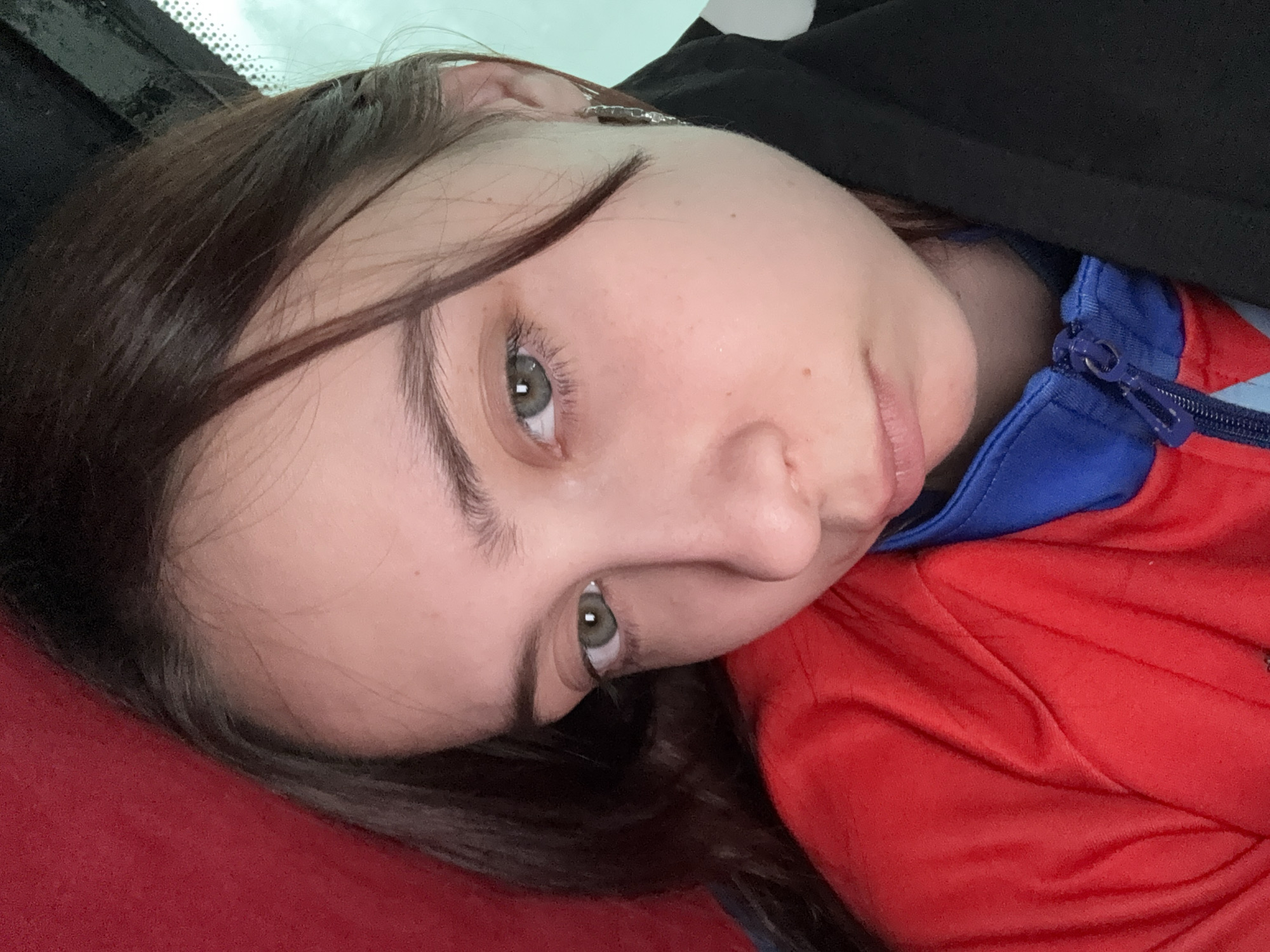 A close-up, angled, self-portrait depicts a young woman. Her face is pale with light brown hair. Her eyes are light green. She is wearing a red jacket with a blue collar. A black garment is draped over her shoulder. A vehicle windshield is visible at the top left of the frame. The setting is likely indoors, possibly inside a car. The location is unknown.