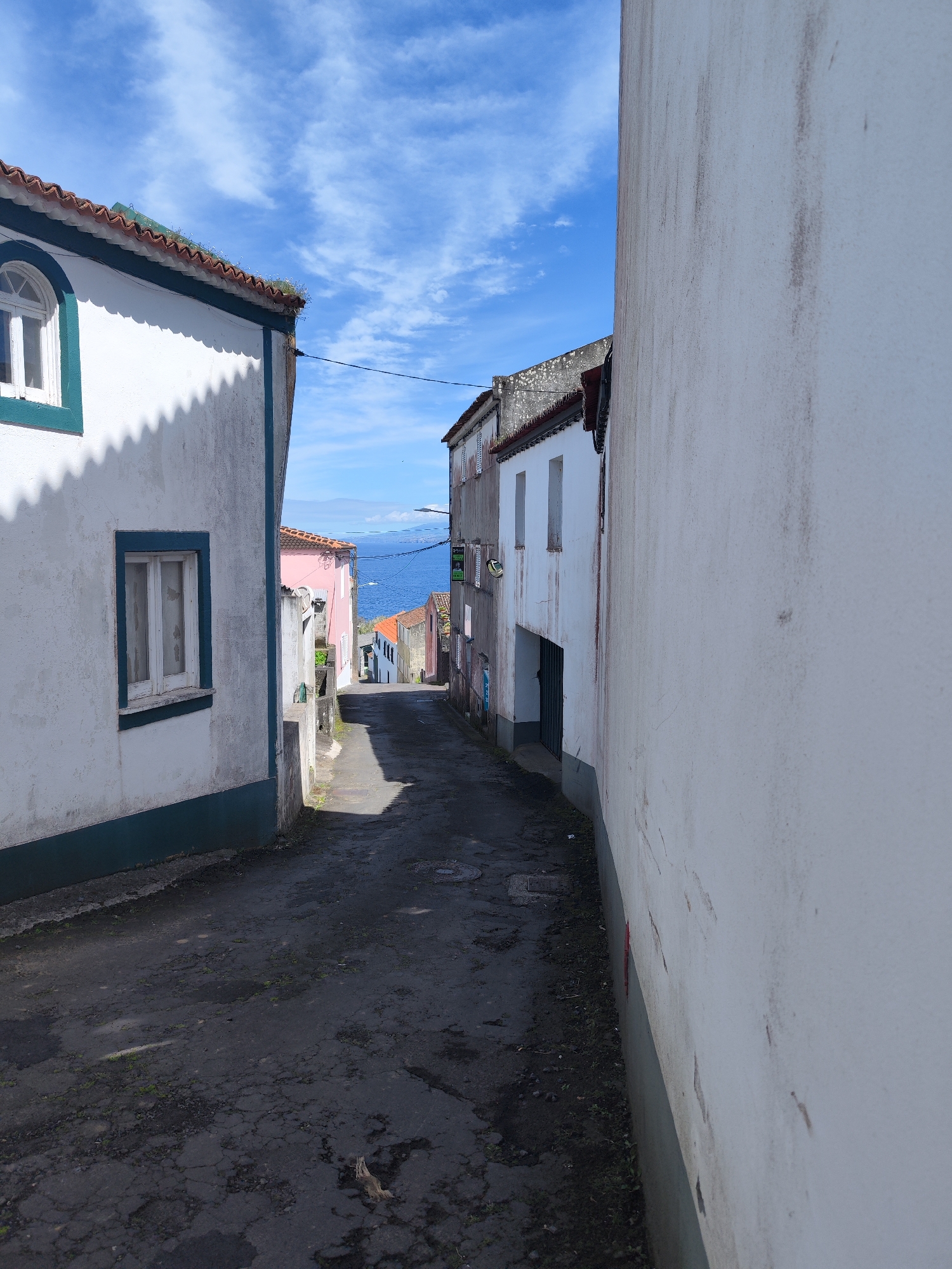 A narrow, paved street, characteristic of a coastal town, descends steeply towards the sea under a bright, partly cloudy blue sky. Buildings with white-washed walls and red-tiled roofs line both sides of the alleyway.
On the left, a prominent building features a distinctive arched window on its upper facade and a rectangular window below, both framed in dark teal or green trim, which also runs along the base of the wall. Greenery sprouts from the edge of its tiled roof, and the building casts a scalloped shadow onto the street from its eaves. The walls show signs of age, with some discoloration and textures.
On the right, a tall, plain white wall dominates the immediate foreground, also showing streaks and signs of wear. Further down, more buildings with similar architectural styles are visible, one featuring a dark green gate or entrance. The street itself is dark, possibly asphalt, with visible wear, cracks, and small patches of moss or weeds.
At the end of the street, a vast expanse of blue ocean meets the horizon, with distant land masses faintly visible. The clear blue sky with scattered wispy clouds suggests a sunny day, likely mid-morning or late afternoon given the shadows. A dark, illegible sign is affixed to a building on the right side further down the street. The scene is quiet and devoid of people, conveying a peaceful atmosphere in what appears to be a historic part of a town like Ponta Delgada, Portugal.