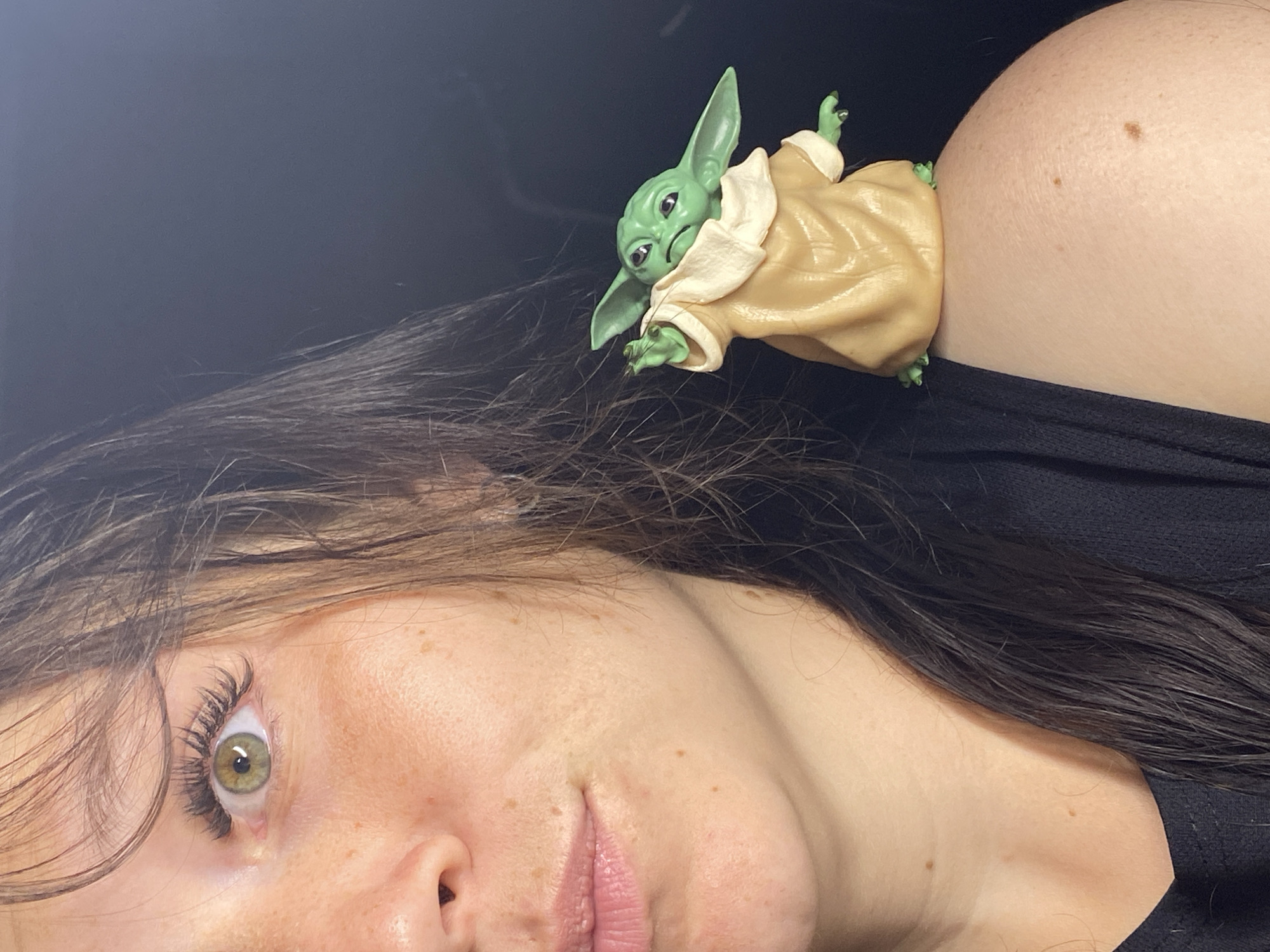 A close-up shot features a woman's face and shoulder with a small, green figurine of the character "Grogu" (also known as Baby Yoda) perched on her shoulder. The woman's left eye, with green irises and long eyelashes, is in focus, as are her freckled cheek and lips. Her dark hair falls around her face and shoulder. Grogu, wearing its signature tan robe, is positioned on the woman's right shoulder, its head tilted and one hand raised as if gesturing. The background is a dark, indistinct blur. The image does not provide any information about the location in the city of Chaikovsky, Russia.