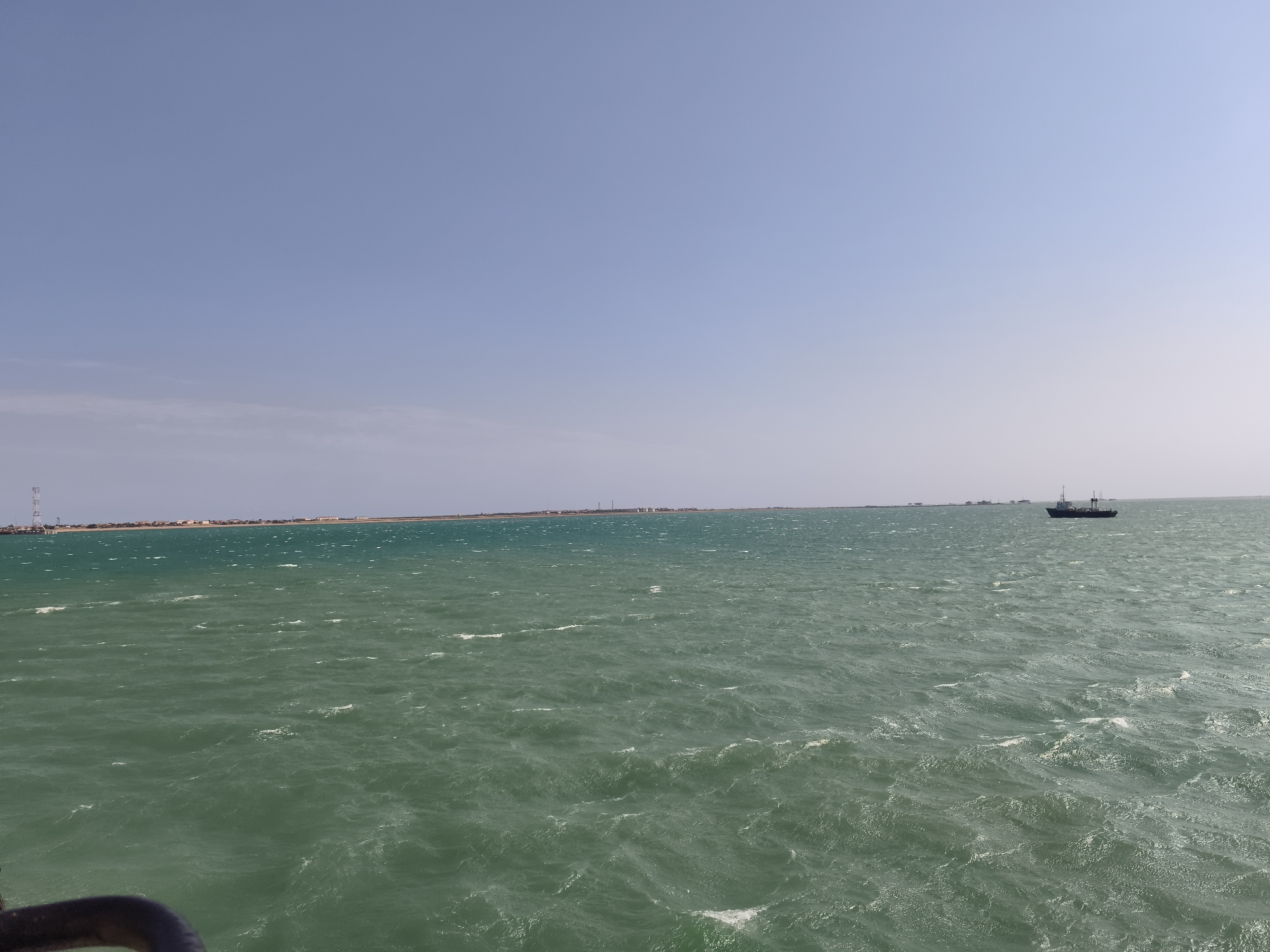 A wide, horizontal shot captures a turbulent, green sea stretching towards a distant, flat coastline under a clear, pale blue sky. The water is choppy, with whitecaps visible across its surface. On the far right of the frame, a dark-hulled ship with a white superstructure sails on the water. In the distance, along the coastline, a thin line of buildings and structures, including a tall tower, is visible. The foreground is partially obscured by a dark, curved object, possibly part of a railing. The scene suggests an open expanse of water, likely a bay or sea, with signs of industrial or coastal development in the distance. This location is consistent with the coastal areas of Pirallahi Raion, Azerbaijan, known for its oil and gas industry and proximity to the Caspian Sea.
