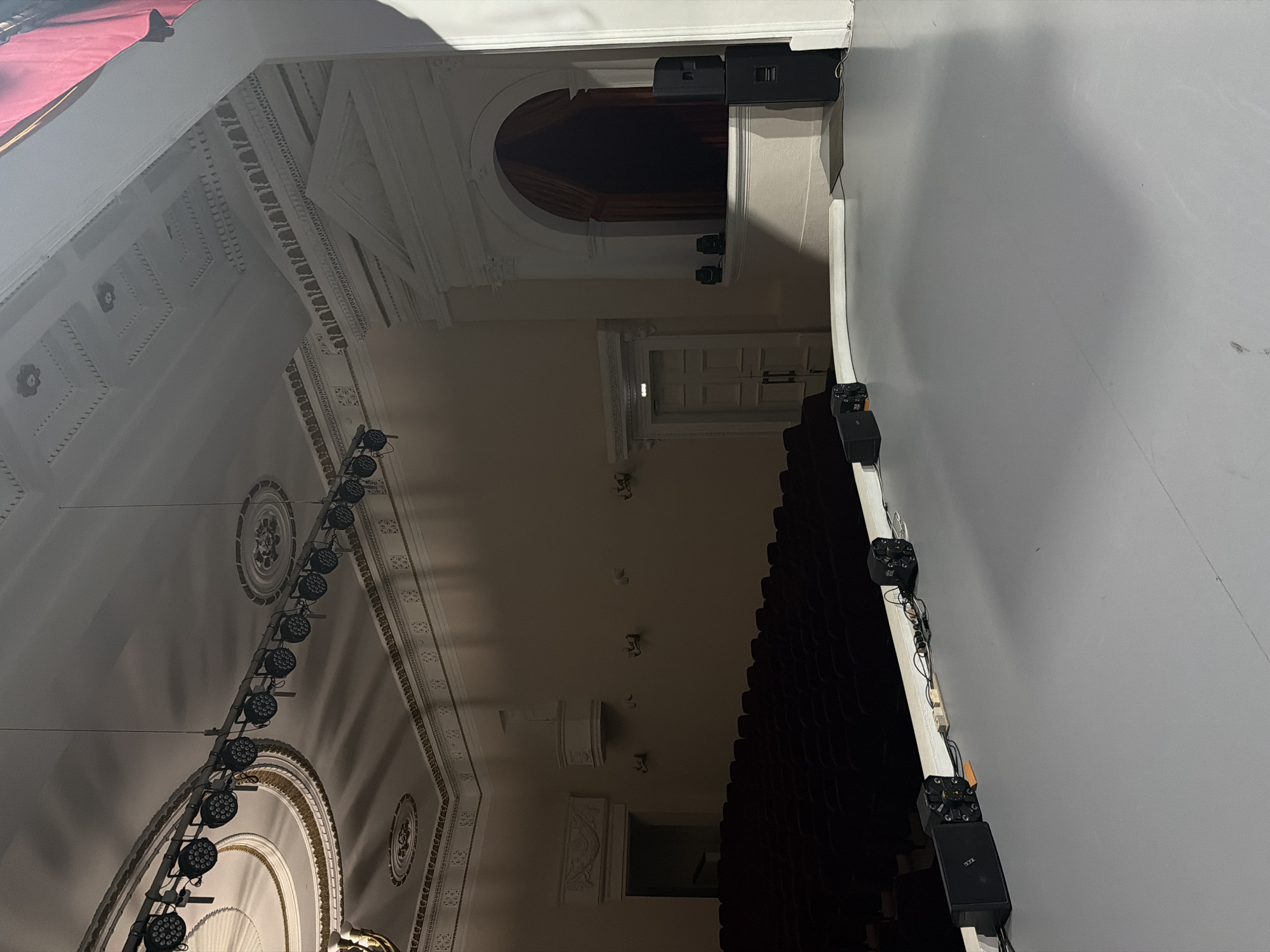 A dimly lit, rotated interior shot appears to be from an auditorium or theater in Perm, Russia. The perspective is from the side of the seating area, looking towards the stage. The left side of the image shows ornate architectural details: a ceiling with decorative moldings, a series of hanging stage lights, and a large arched structure. The center shows the seating area with rows of red seats. The right side depicts a white wall with electrical boxes and wiring. A doorway can be seen at the back of the auditorium, located above the seating area.