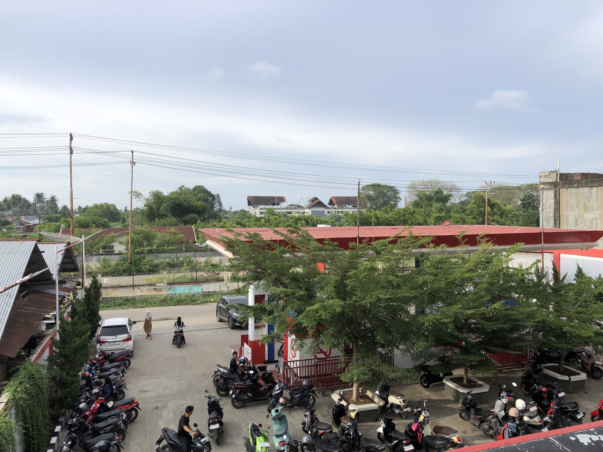 The image captures an outdoor scene at a gas station in Geulumpangsulu Timu, Indonesia. The time of day appears to be daytime, with overcast skies suggesting potential for rain or a generally cloudy atmosphere.
The foreground is dominated by numerous parked motorcycles, a common mode of transportation in the region. Several people are visible, some riding motorcycles and others standing or walking near them. Two individuals are walking near a white car parked in a lot. A person is seated on a motorcycle at the gas pumps, and another is standing near a motorcycle. Further back, several more individuals are either walking or on their bikes.
The gas station itself is characterized by a prominent red roof structure, typical of such facilities. Trees with lush green foliage are scattered throughout the scene, some planted in rectangular planters. In the background, buildings with red roofs and a large concrete structure are visible, along with a dense line of trees and vegetation, suggesting a semi-urban or suburban environment. Overhead power lines with wooden poles crisscross the sky, a common sight in many Indonesian locales.
A white car with an Indonesian license plate (likely reading "BL 1183 Y") is parked in the vicinity of the gas station, and a dark-colored SUV is positioned near the pumps. The overall impression is one of a bustling, everyday scene at a local service station. No specific emotions are discernible from the image; the focus is on the activities and objects present in the setting.