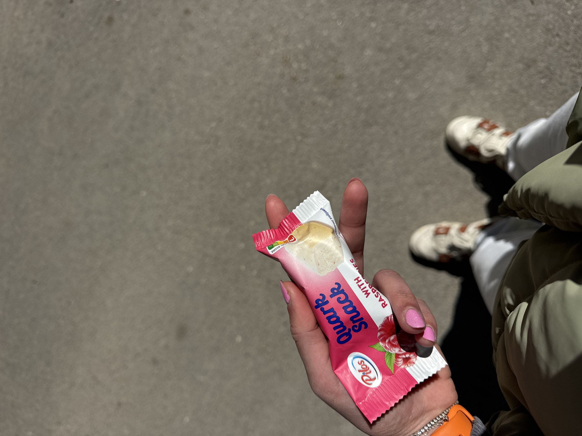 The image shows a person holding a "Quark Snack with Raspberries" bar, branded "Plus." The bar is pink and white with an image of a raspberry. The person's hand has pink nail polish, and they are wearing a silver bracelet and an orange watch. The lower part of the image shows parts of the person's outfit: white pants, white and brown sneakers, and a green jacket. The background appears to be an outdoor setting, likely a paved street. Considering the presence of the "Plus" brand food product, which is available in Bulgarian markets, the location could likely be in Sofia, Bulgaria.