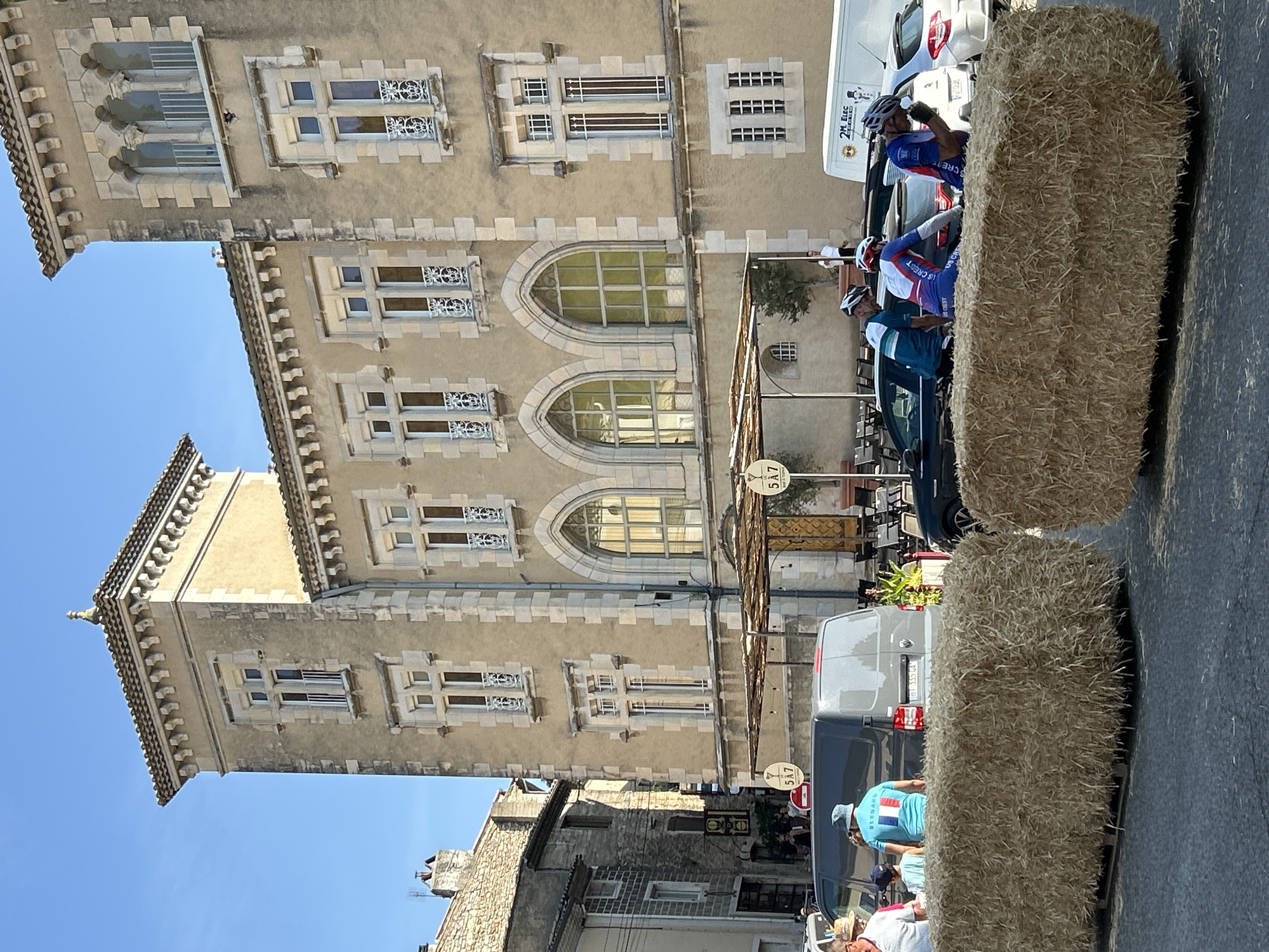 This is a street scene in the city of Marsanne, France, featuring a building with distinctive architecture, likely dating from an earlier period, judging by the stone facade and arched windows. In the foreground, two large bales of hay are placed on the asphalt road, acting as a temporary barrier. Three cyclists, dressed in racing attire, are seen in the middle ground, appearing to be either resting or preparing for an event. One cyclist is wearing a blue and white striped jersey, another a blue, white, and red patterned jersey, and the third a blue jersey with red accents. Behind the hay bales and cyclists, a grey van is parked. Further back, part of a building with a tiled roof and stone walls is visible, suggesting a historic district. A few people are standing near the van. The scene is bathed in bright sunlight, casting shadows and indicating a clear day. The building in the background has multiple stories with numerous windows, some of which are decorated with ornate ironwork. A covered outdoor seating area, possibly a cafe or restaurant, is situated in front of the building. The overall impression is of a town square or street that has been prepared for a public event, such as a cycling race.