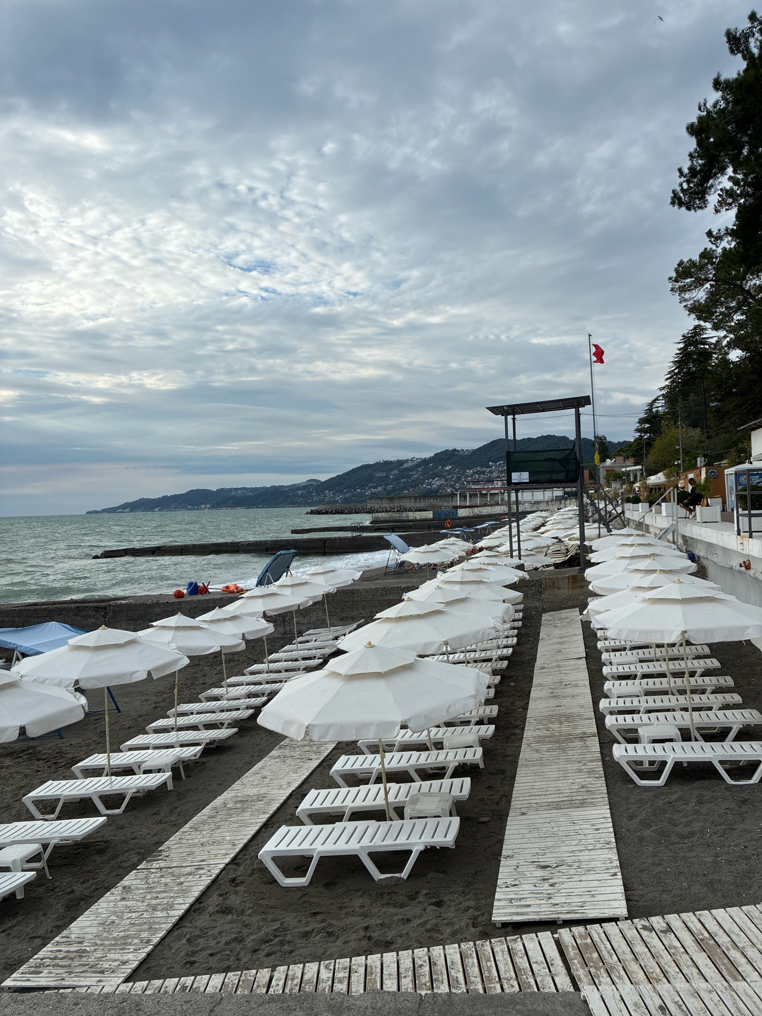 This image depicts a beach in Sochi, Russia, under a cloudy sky. The foreground is dominated by rows of white lounge chairs and umbrellas, arranged on dark sand. Wooden walkways are laid out between the rows of chairs, providing access to the beach. In the background, the sea stretches out to the horizon, with a gentle wave action. Beyond the sea, a hilly coastline is visible, lined with buildings. A lifeguard tower stands prominently on the right side of the beach, topped with a red flag. A few people can be seen in the distance, some seated near a building on the right. The overall atmosphere is calm and somewhat subdued due to the overcast weather.