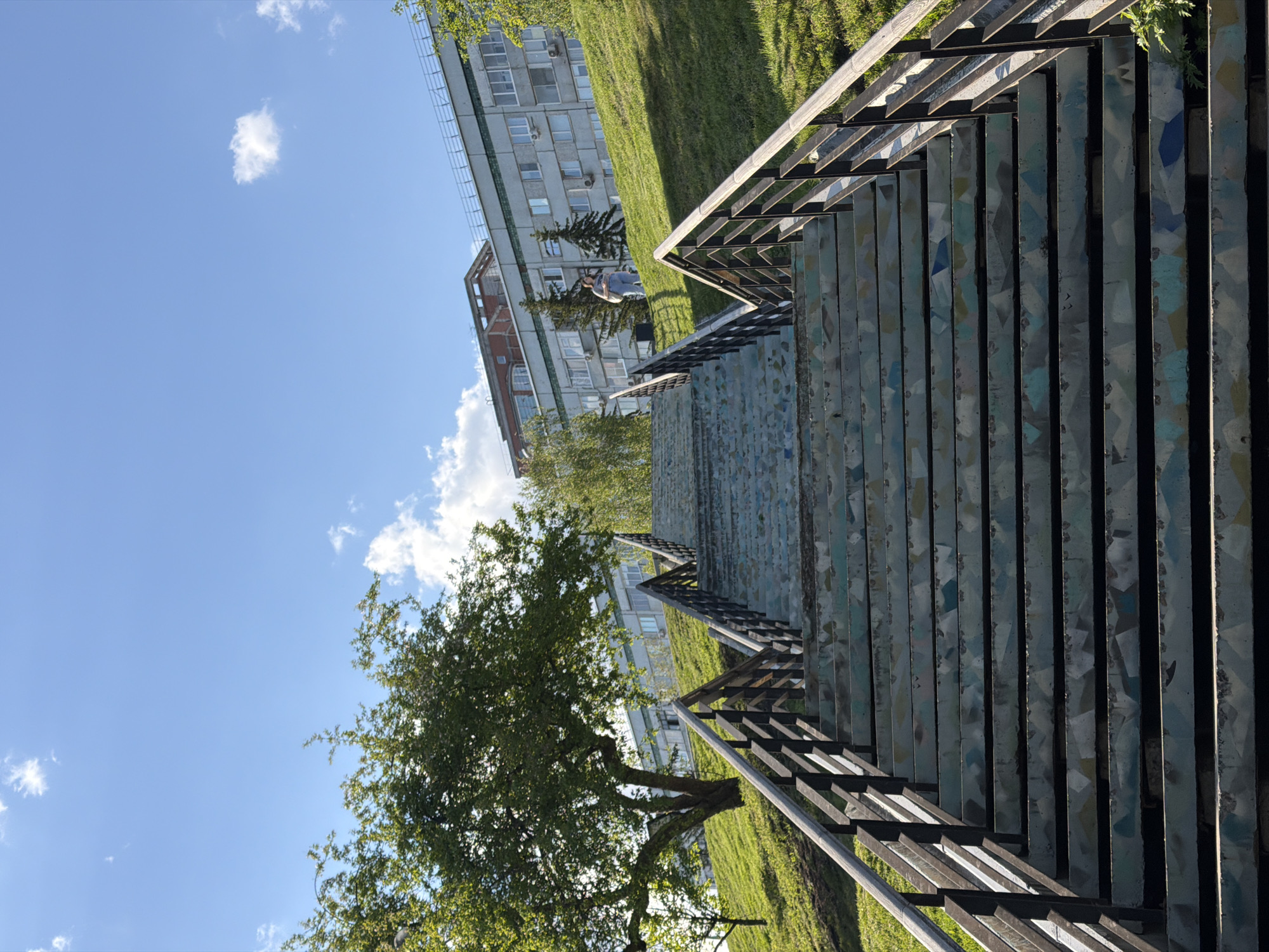 The image shows a view looking down a long staircase with metal railings on both sides. The staircase steps appear to be made of patterned tiles. In the background, a building is visible, along with grassy areas and trees. The sky is blue with some clouds. A person is walking near the building in the background. The location is the mosaic stairs in Krasnoyarsk, Russia.