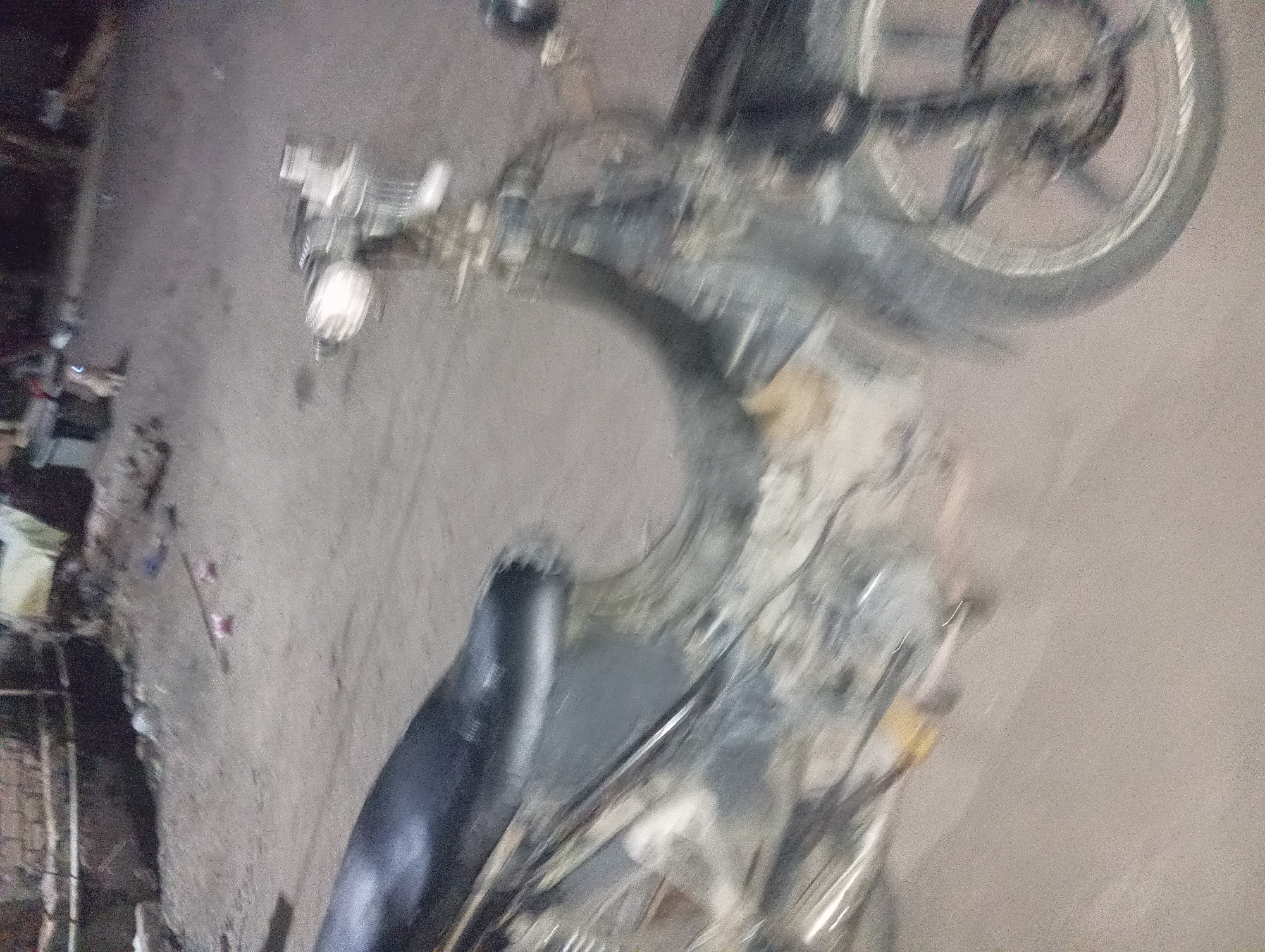 The image depicts a blurry, motion-affected scene, likely taken at night or in low light conditions, in the city of Rejosari, Indonesia. The primary subject is a black motorcycle, angled to the right, with its front wheel and handlebars visible. The motorcycle appears to be in motion, as indicated by the motion blur across the entire image.
The background is indistinct due to the blur, but it suggests an outdoor urban or semi-urban environment. Hints of structures, possibly buildings or walls, are visible in the upper left portion of the image. The ground appears to be a dirt or gravel surface. There are small, indistinct objects on the ground, possibly debris or litter.
In the upper center of the image, there is a faint, blurred shape that could be a small animal, possibly a dog, standing on the ground. The overall impression is a fleeting moment captured while the motorcycle was moving through a somewhat unkempt area in Rejosari.