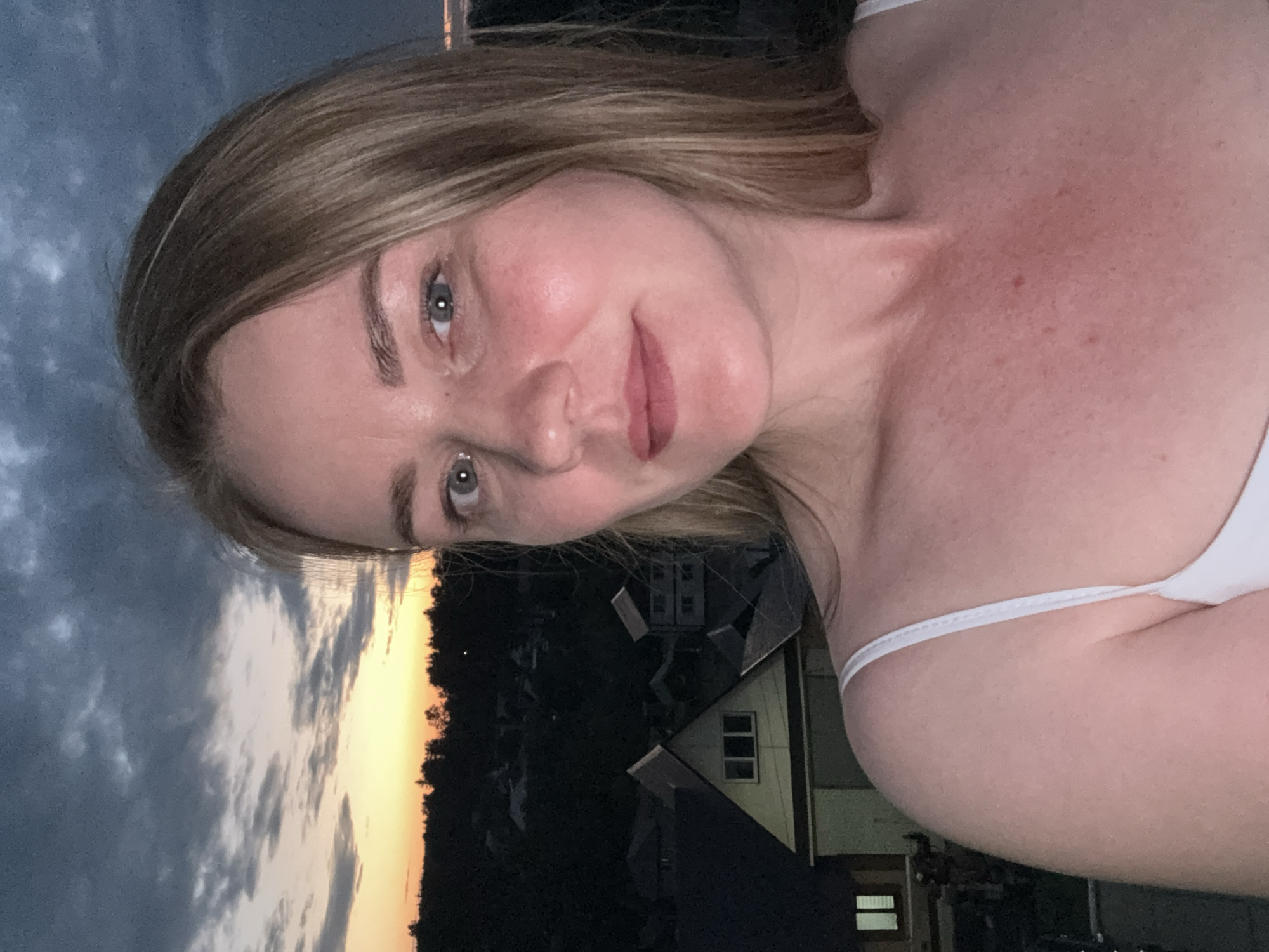 A close-up selfie of a woman with blonde hair and blue eyes is taken outdoors at dusk. She is wearing a white tank top. Behind her, the sky is a mix of dark clouds and a bright orange sunset. Below the sky, a dark silhouette of trees is visible, with houses and rooftops of a village nestled in the landscape. The scene appears to be in a rural or suburban area, possibly on the outskirts of Chaikovsky, Russia, given the architecture of the houses and the prevalent twilight conditions. The overall mood is serene, with the natural light of the setting sun creating a soft glow.