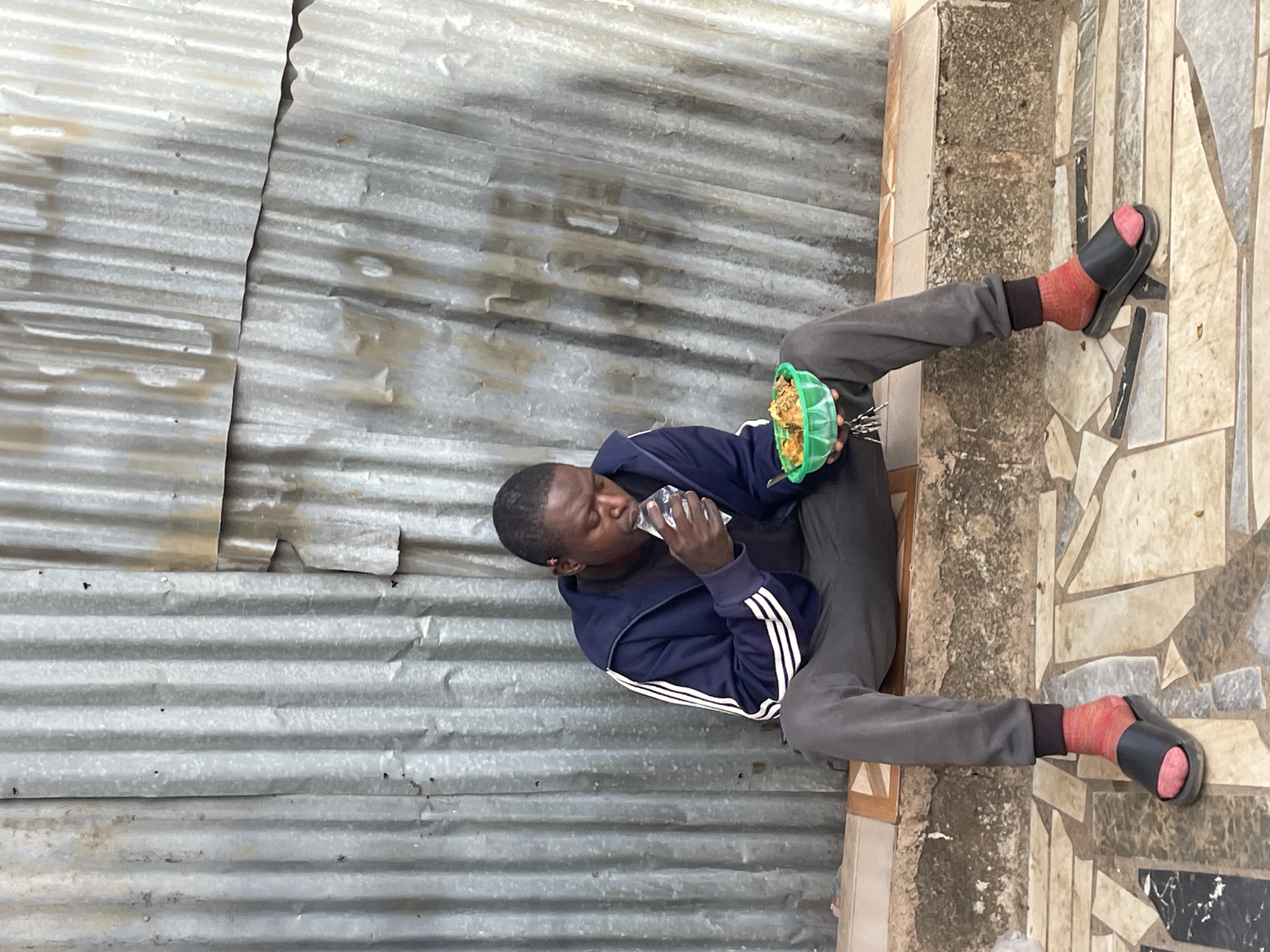 This image, captured in Jos, Nigeria, features a man seated on the ground and consuming a meal. He is positioned against a weathered wall constructed from corrugated galvanized iron sheets, which are characterized by horizontal ridges and a visible vertical seam.
The man has dark skin and short, dark hair. He is dressed in a navy blue track jacket featuring white stripes on the sleeves, a dark shirt underneath, and grey track pants. His feet are covered with bright pink or red socks, and he wears black slide sandals.
In his left hand, the man holds a small, light green, possibly plastic, bowl filled with a meal that appears to be a mix of grains or noodles with other ingredients. With his right hand, he holds a clear plastic sachet or bag to his mouth, suggesting he is drinking from it, likely water or another beverage. His facial expression is neutral and focused on the act of eating and drinking.
The ground beneath him is a tiled surface, displaying an irregular mosaic pattern in shades of beige, brown, grey, and black. A rough-textured concrete or stone strip forms a boundary between the tiled floor and the corrugated metal wall. The natural lighting indicates it is daytime. There is no visible text in the image.