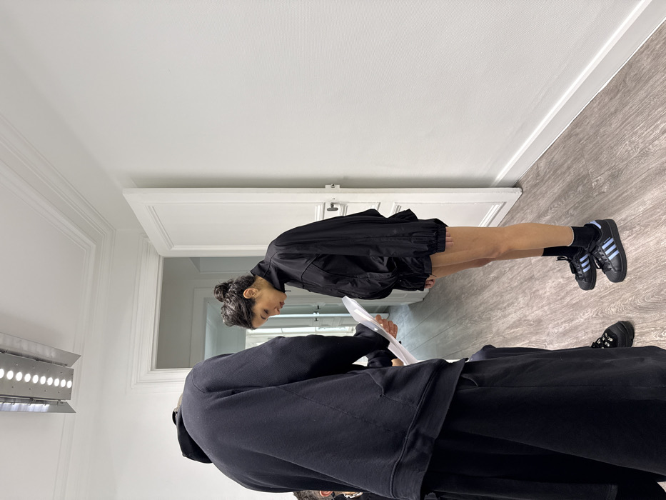The scene depicts an interior space in Paris, France, characterized by white walls with crown molding near the ceiling and a wood-patterned floor. A partially open white paneled door is visible in the background. Overhead, a rectangular LED lighting fixture is mounted on the ceiling.
Two individuals are present. In the foreground, facing away from the camera, is a person wearing an oversized black hooded sweatshirt and what appear to be baggy black pants or a skirt, with thick-soled black shoes on their left foot. This individual holds a translucent sheet or document in both hands, extended towards the second person.
The second individual, positioned in profile facing left in the mid-ground, has dark hair styled in a bun and is looking downwards. They are wearing a black high-collared, long-sleeved jacket that appears oversized and cinched at the waist. Their bare legs are visible from mid-thigh down. They wear black ankle socks and black Adidas Superstar sneakers featuring three light blue stripes and a black shell toe.
The interaction suggests a review or presentation, with one person holding a document for the other, who appears to be observing or listening attentively.
