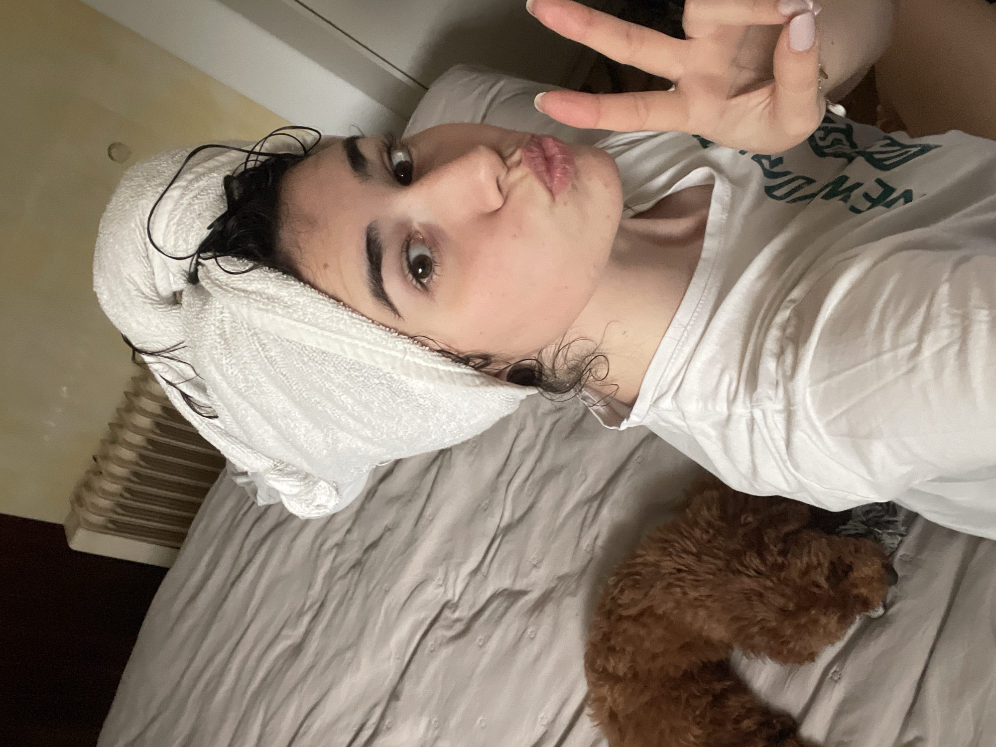 A young woman is captured in a selfie, lying on her back on a bed. Her head is wrapped in a white towel, with wet, dark hair visible around the edges. She looks directly at the camera with large, dark eyes, making a "kiss face" with puckered lips. Her right hand is raised, showing a "peace sign," and her fingernails are painted a light, neutral color.
She is wearing a white t-shirt with green text, partially visible as "NEW YOR," suggesting "NEW YORK." Beside her on the light grey or beige duvet is a small, fluffy brown dog, possibly a poodle or doodle, resting. In the background, to the left, a light brown radiator with horizontal fins is visible against a light-colored wall, indicating an indoor setting, likely a bedroom. The overall scene appears relaxed and playful, suggesting the photo was taken after a shower or bath. The radiator design is consistent with architecture found in Sofia, Bulgaria.