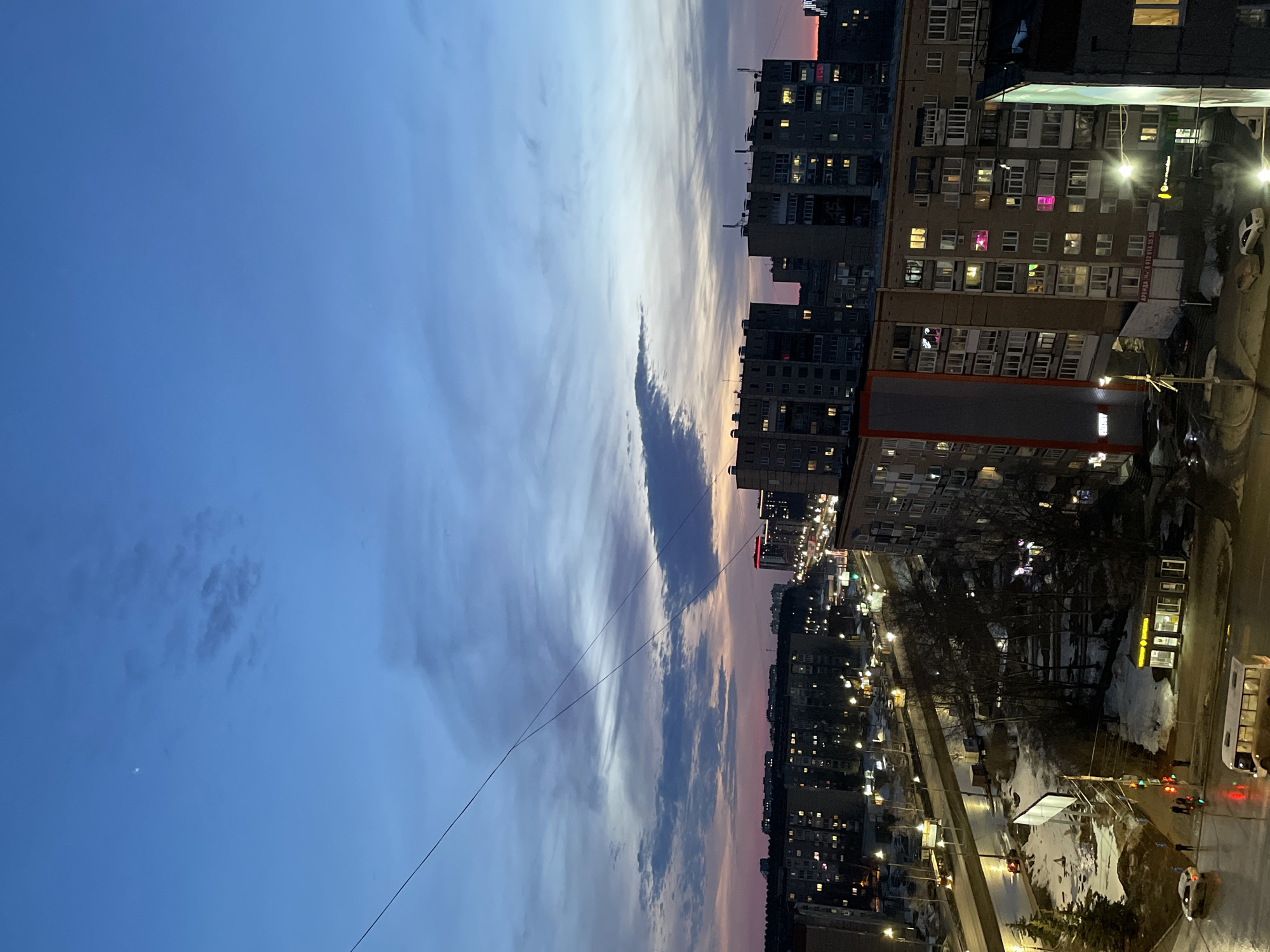 The image depicts an urban cityscape in Izhevsk, Russia, captured from an elevated perspective during twilight or early evening. The sky, transitioning from deep blue overhead to hues of purple, orange, and pink near the horizon, contains scattered clouds. A faint crescent moon is visible in the upper left quadrant. Multiple utility wires or power lines diagonally traverse the upper half of the frame.
The foreground and midground feature a dense arrangement of multi-story residential or commercial buildings, predominantly grey and beige, with numerous windows illuminated in yellow, white, pink, and red. One prominent building exhibits a grey and orange facade with "АРЕНДА" (Rent) signs. Below these structures, a network of illuminated streets and roads is visible. The ground is characterized by patches of melting snow and slush, with wet pavement reflecting streetlights, suggesting a late winter or early spring setting. Bare deciduous trees are interspersed among the buildings and along the roadsides.
Several vehicles are present on the roads; a white bus is in the immediate foreground, and various cars, some with active headlights or taillights, are visible in motion and parked. Streetlights cast bright, yellow-white illumination onto the scene. At least one traffic light is active, displaying both red and green signals. Pedestrians are observed on the sidewalks near street crossings, one group of four individuals near a red traffic light and another individual further down the street. A small, well-lit commercial kiosk with "ТАБАК" (Tobacco) visible on its signage stands near the sidewalk. Large, unlit billboards are also present on the ground level. In the distance, additional buildings with illuminated windows extend towards the horizon, where distant city lights are visible.