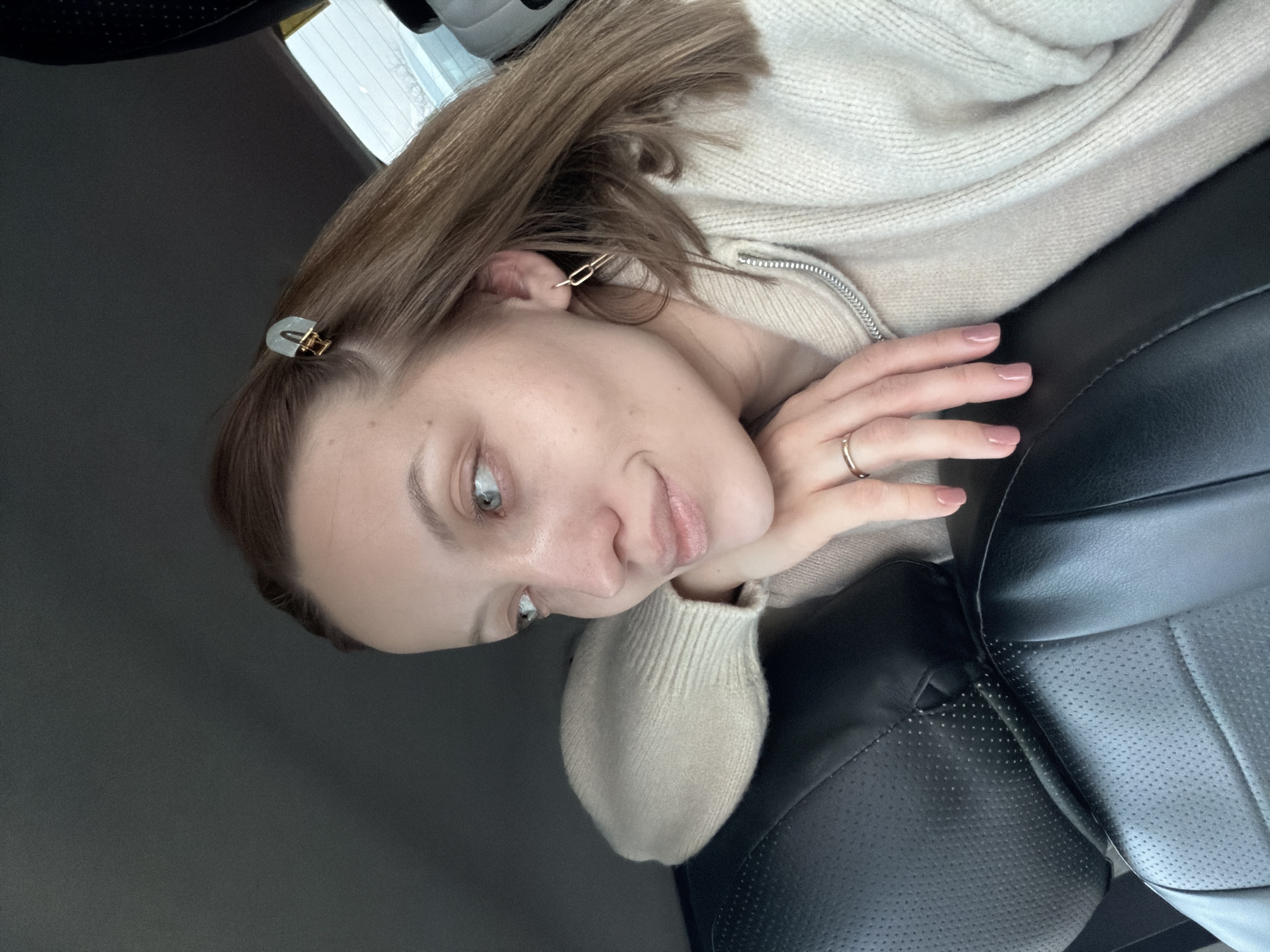 A light-skinned woman with fair brown hair is in a car. She wears a light beige sweater and has hair clips in her hair. Her fingernails are painted a light pink. A gold ring is on her ring finger. The car has black leather seats. The location of the image cannot be determined to be Reshetnikovo, Russia from the image alone.