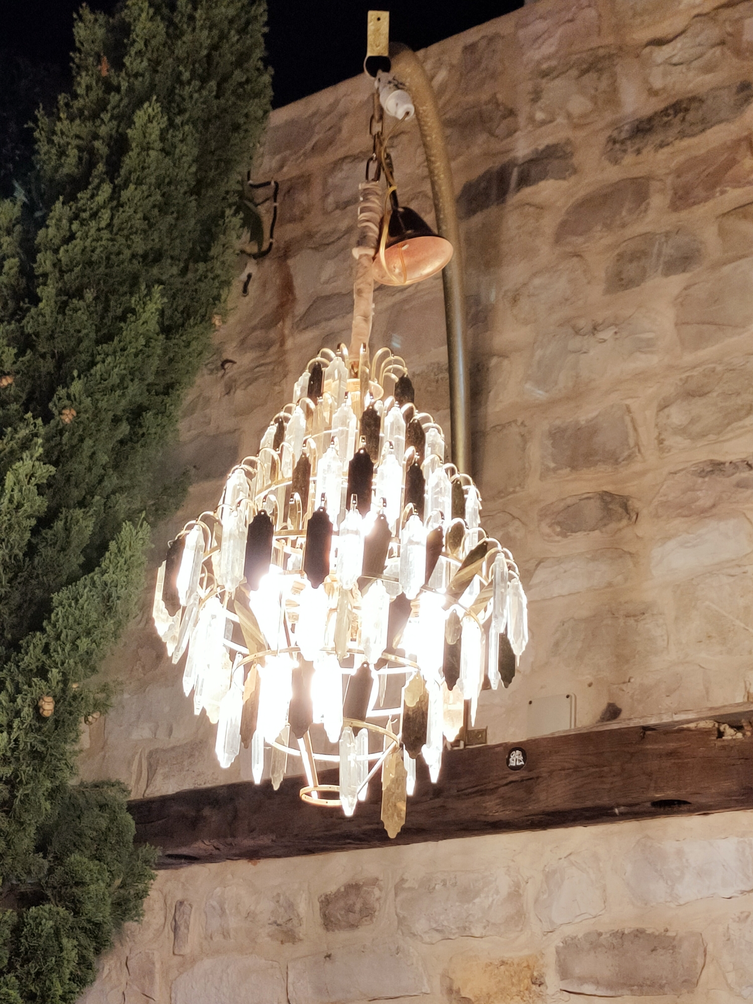 This is a medium shot, eye-level, indoor photograph of a decorative chandelier with clear and dark crystal-like prisms, hanging in front of a stone wall. The chandelier is gold-toned and appears to be lit from within, illuminating the prisms. To the left of the chandelier is a tall, green evergreen tree. A thick, dark wooden beam is visible horizontally across the lower portion of the image, resting on the stone wall. The stone wall is constructed from irregularly shaped, light-colored stones. The scene appears to be outdoors, potentially in an area with a rustic or historic architectural style, possibly in Cebalat, Tunisia, given the stone construction and the presence of an evergreen tree which are common in Mediterranean climates. There are no people or animals visible in the image.