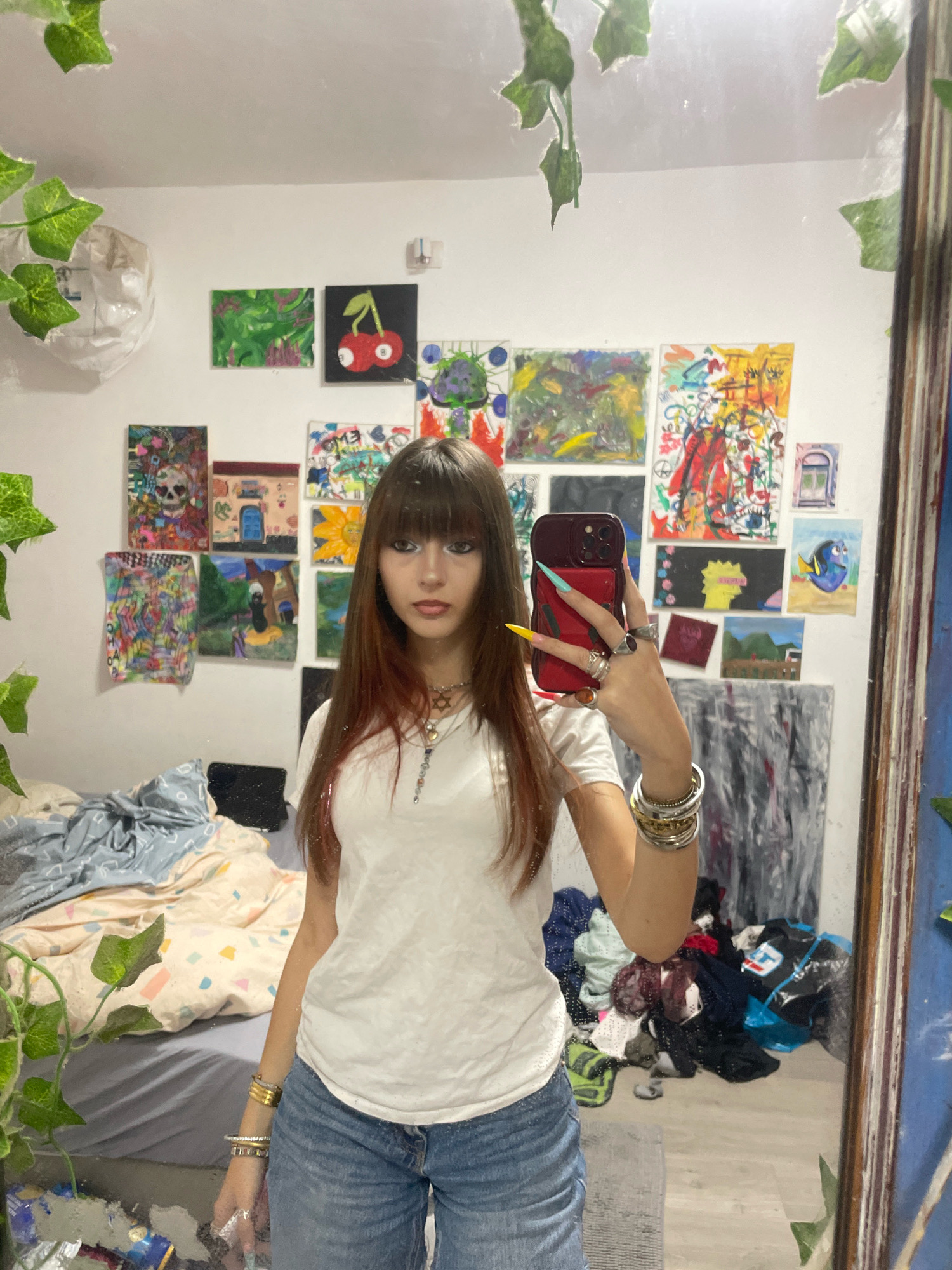 The image depicts a young woman taking a mirror selfie in what appears to be a bedroom. The room is decorated with numerous small, colorful paintings adorning the walls, featuring a variety of subjects including abstract designs, a skull, a house, a sunflower, and cartoon characters. To the left of the woman, a bed is visible with rumpled bedding. A pile of laundry is also present in the room, reflected in the mirror. The woman is wearing a white t-shirt and blue denim jeans, and has long brown hair with reddish highlights. She is wearing several rings and bracelets on her left hand, which is holding up a red smartphone to take the picture. The overall impression is of a personal, artistic space, likely a bedroom or a personal studio. This scene is located in Raanana, Israel.